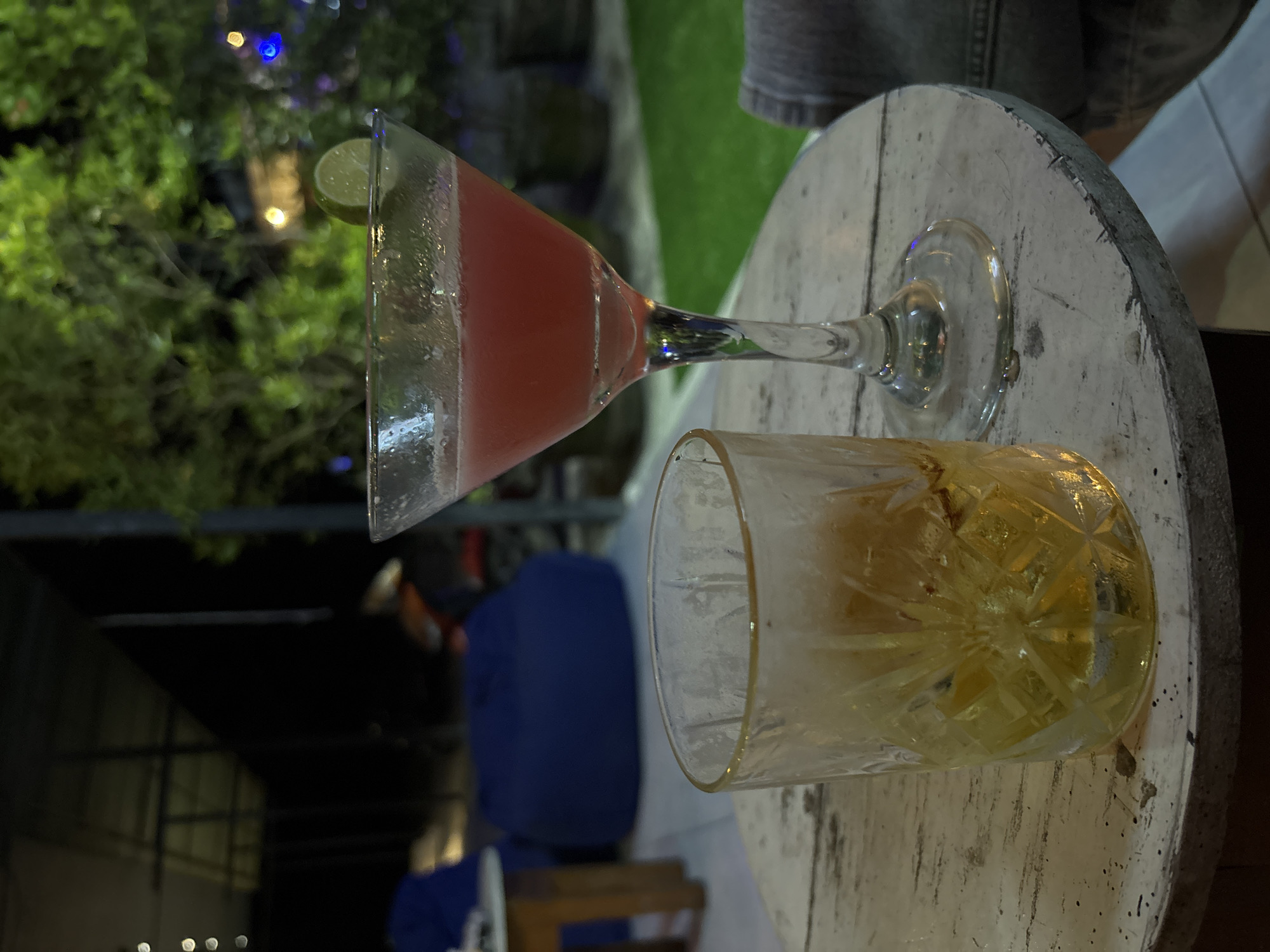 A horizontally oriented, slightly blurry, medium-angle shot depicts two partially consumed alcoholic beverages resting on a small, round, weathered white table with a metal band around its edge. A pink martini with a lime wedge on the rim sits in a clear glass with a long stem. Beside it, a clear lowball glass holds a yellow liquid, likely an alcoholic drink like whiskey or a mixed drink, with a patterned design visible through the liquid. The table appears to be outdoors, as suggested by the greenery and string lights visible in the background. The setting is night. A person wearing a denim jacket sits at the table. The location is likely in Kuta Selatan, Indonesia, though there are no specific elements to confirm it.