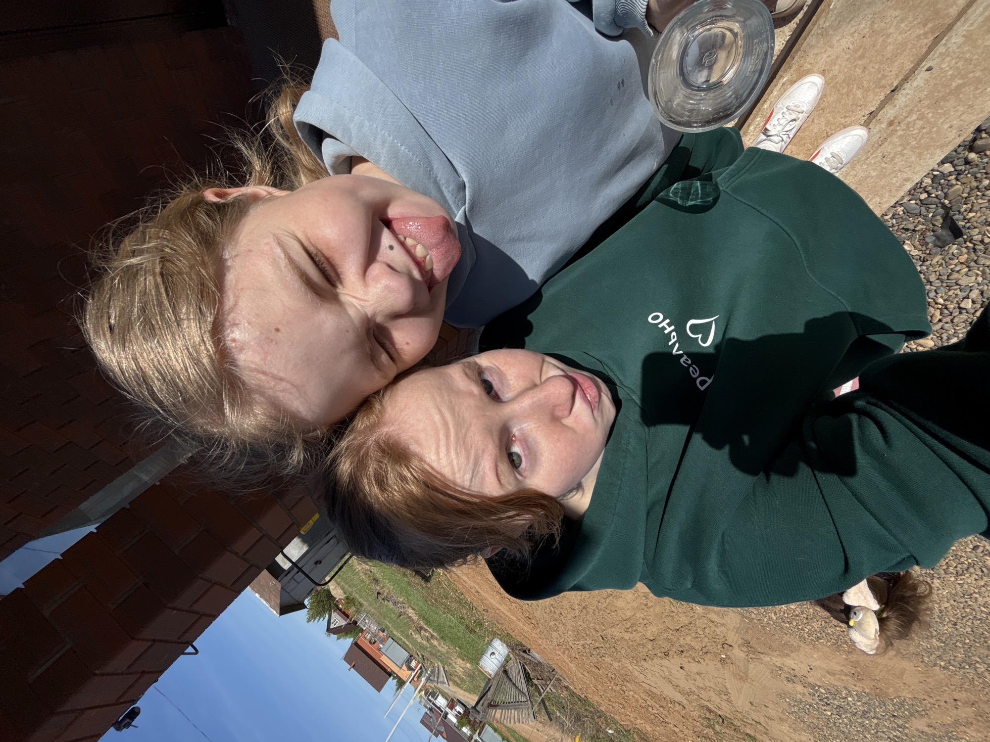 Two young individuals are lying side-by-side outdoors. The person on the left has blonde hair and is sticking out their tongue. They are wearing a light blue hooded sweatshirt. The person on the right has reddish-brown hair and is wearing a dark green hooded sweatshirt with lettering in Russian "Реально" and a heart. A clear glass dish is partially visible above them on what appears to be a weathered wooden surface. White sneakers with red accents are nearby. The background shows a rural landscape with houses and a blue sky, suggestive of a location in the city of Prikamskii, Russia. The ground is a mix of dirt and small stones. A brick structure is partially visible on the left side. The photograph is taken from a slightly elevated perspective.