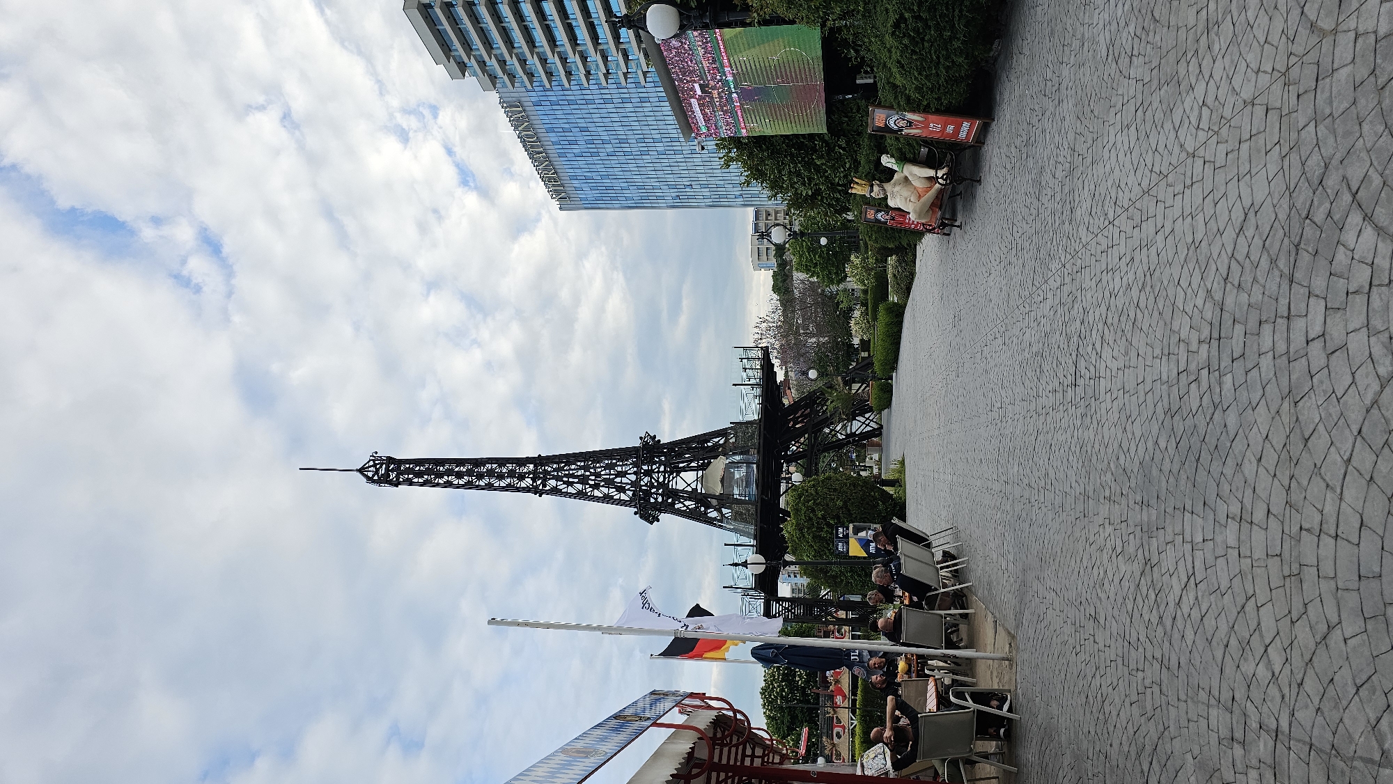This image shows a replica of the Eiffel Tower in Varna, Bulgaria. The tower is located in a park-like setting with a paved walkway. In the foreground, there is an outdoor seating area with people sitting at tables. There is a German flag visible along with flags with text. In the background is a modern building, a screen showing a sports game, and a sculpture on a bench. The sky is partly cloudy.