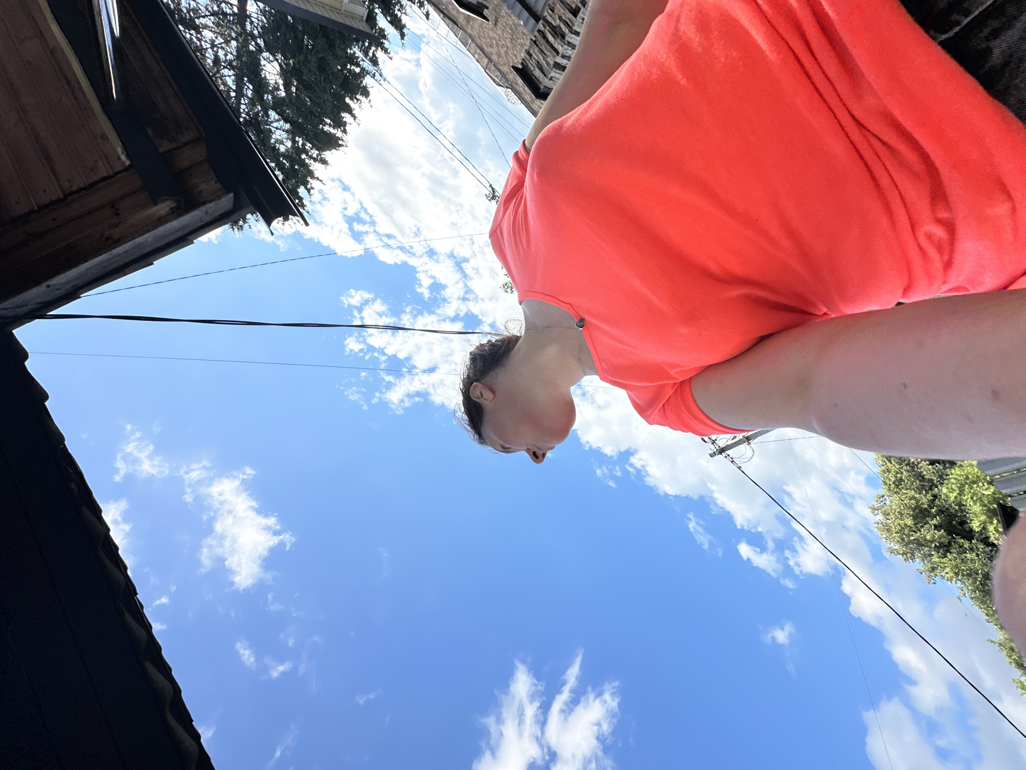 The image features a person wearing a bright orange t-shirt, seen from a low-angle perspective, looking upwards into a bright blue sky with scattered white clouds. The person's head and upper body are visible. In the background, there's a glimpse of buildings and trees. A dark-colored roofline is visible in the lower left corner. Electrical wires are also visible stretching across the sky. The lighting suggests it is daytime, likely a clear, sunny day. The overall mood seems relaxed and observational, as if the person is enjoying the sky. The location is identified as Nizhny Novgorod, Russia. No visible text is present in the image.