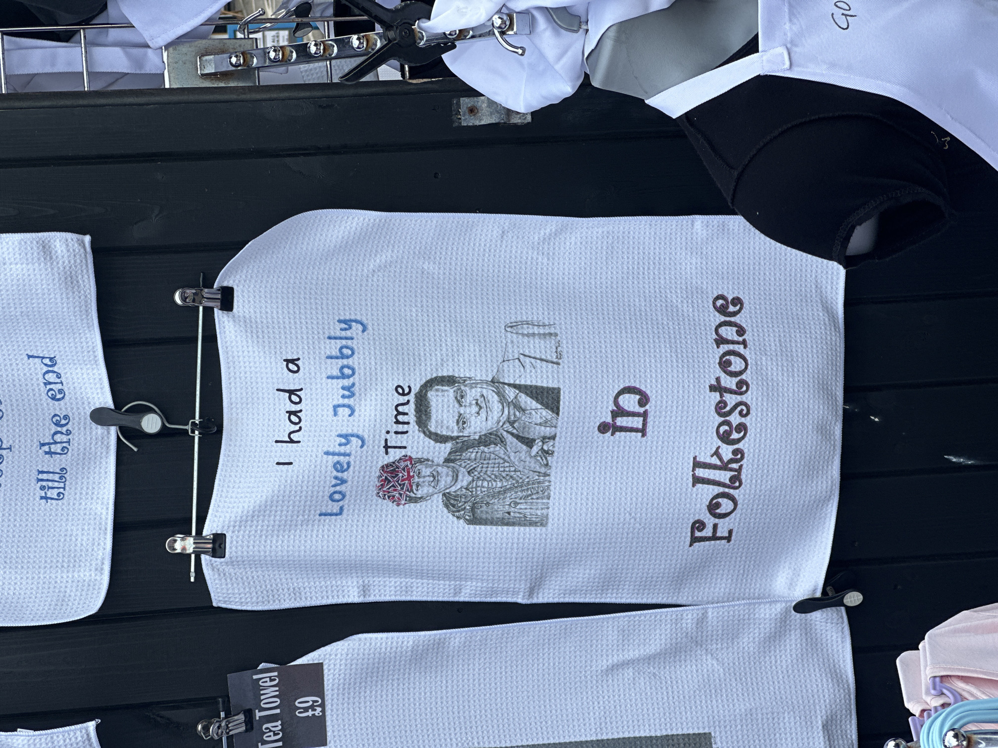 The image shows a white tea towel hanging on a display rack, likely for sale, adorned with the phrase "I had a Lovely Jubbly Time in Folkestone" printed on it. Below the phrase, there's a sketched image of two characters, possibly the iconic British comedy duo Del Boy and Rodney Trotter, known for their catchphrase "Lovely Jubbly" from the TV series Only Fools and Horses.
The tea towel is hanging alongside others, as evidenced by the "Tea Towel £9" tag visible beneath. The setting appears to be a shop or market stall, given the display racks. The overall tone suggests a humorous and nostalgic souvenir, referencing British popular culture and the seaside town of Folkestone, located in the Folkestone and Hythe District, United Kingdom.