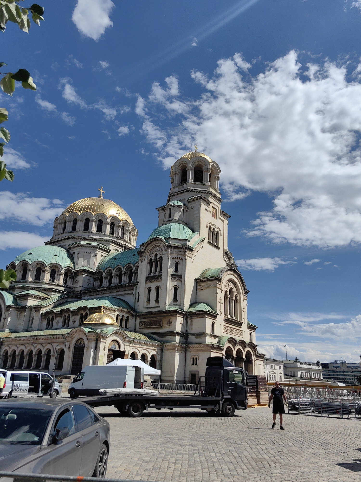 This is a photograph taken outdoors in front of the Alexander Nevsky Cathedral in Sofia, Bulgaria. The cathedral, a large and ornate structure with gold domes and green roofs, dominates the upper portion of the frame against a bright blue sky filled with white, puffy clouds. In the foreground, a cobblestone plaza is visible. Several vehicles are present on the plaza, including a gray sedan in the lower left corner, a white van, and a black flatbed truck. A person wearing a black t-shirt and shorts is standing in the middle of the plaza, facing away from the camera. Various scaffolding and temporary structures are set up in the background, suggesting ongoing events or construction. The lighting indicates it is daytime, likely a sunny afternoon.