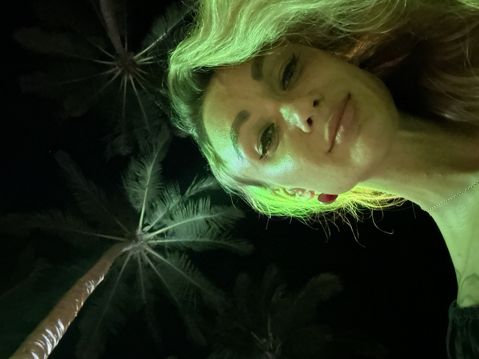 The image captures a nighttime outdoor scene in Mũi Né, Vietnam. The primary subject is a female individual, positioned in the foreground, looking directly into the camera from a low-angle perspective. Her face, neck, and light-colored hair are prominently illuminated by a strong green light source, casting a distinct green hue. She exhibits a subtle smile with slightly parted lips and wears a dark top, a silver-toned chain necklace, and a red heart-shaped earring on her visible left ear. The background consists of multiple palm trees, largely silhouetted against a dark night sky, with the undersides of some fronds also catching the green illumination.