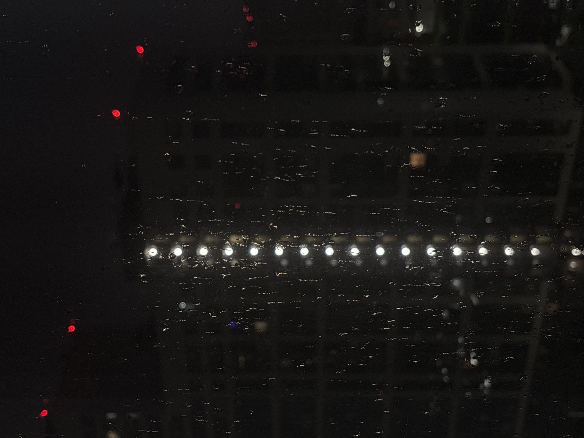 The image depicts a nighttime view through a wet window, likely in the city of Sinyavino, Russia. The scene is dark, with blurred lights from buildings in the background. A vertical line of white circular lights, possibly from a building's illumination, is visible down the center of the frame. Scattered red lights, also blurred, are present in the upper portion of the image. The windowpane is covered in raindrops, creating streaks and distorting the view of the external environment. The overall impression is of a rainy urban night, with the city lights softened and diffused by the water on the glass. There are no people or discernible actions captured in the image.