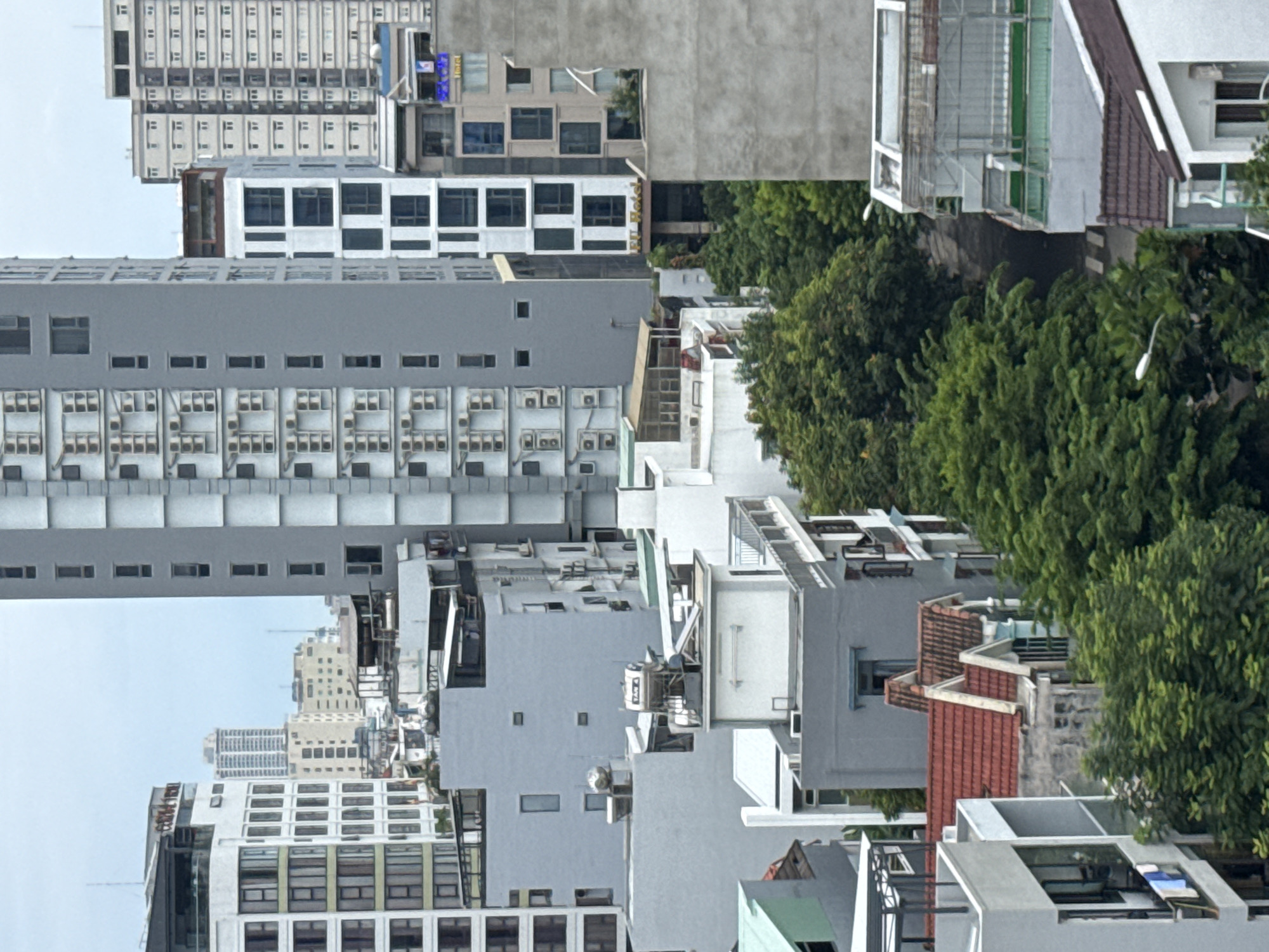 The image shows an urban landscape in Đà Nẵng, Vietnam. Tall buildings dominate the scene, some with multiple air conditioning units. Trees are interspersed among the buildings. There are also rooftops with red tiles. The overall scene is dense and congested. The sky is overcast.