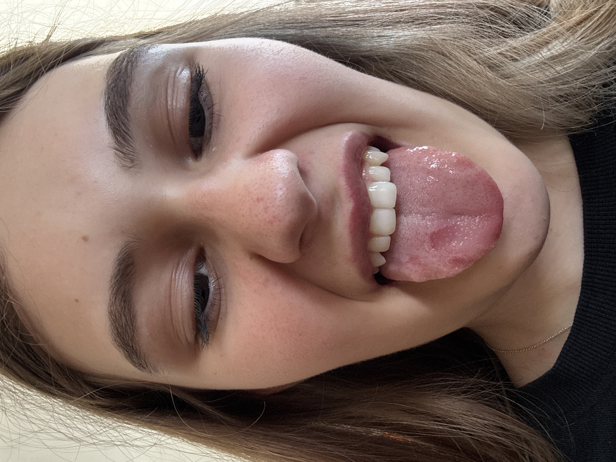 A close-up, vertically-oriented shot captures a person's face tilted sideways, depicting a playful expression. The subject, likely female, has long, light brown hair that frames her face and is visible along the top and bottom edges of the frame. Her skin appears smooth, with some subtle freckles visible on her nose and cheek.
Her eyes are open, with long, dark eyelashes suggesting makeup. Her eyebrows are well-defined and dark. The most prominent feature is her wide-open mouth, from which she extends a healthy, pink tongue. Her front teeth are visible, appearing bright white and uniformly shaped, possibly indicating dental work such as crowns or veneers. The corners of her mouth are turned upwards, suggesting a smile or a mischievous, silly gesture.
A thin, gold-colored chain necklace is visible around her neck, resting against a dark-colored garment, possibly a sweater or shirt. The background is uniformly light-colored and out of focus, providing no specific environmental cues about the location, other than implying an indoor setting. The overall mood conveyed is lighthearted and informal. No visible text is present in the image. The provided context indicates the location is Panagyurishte, Bulgaria.