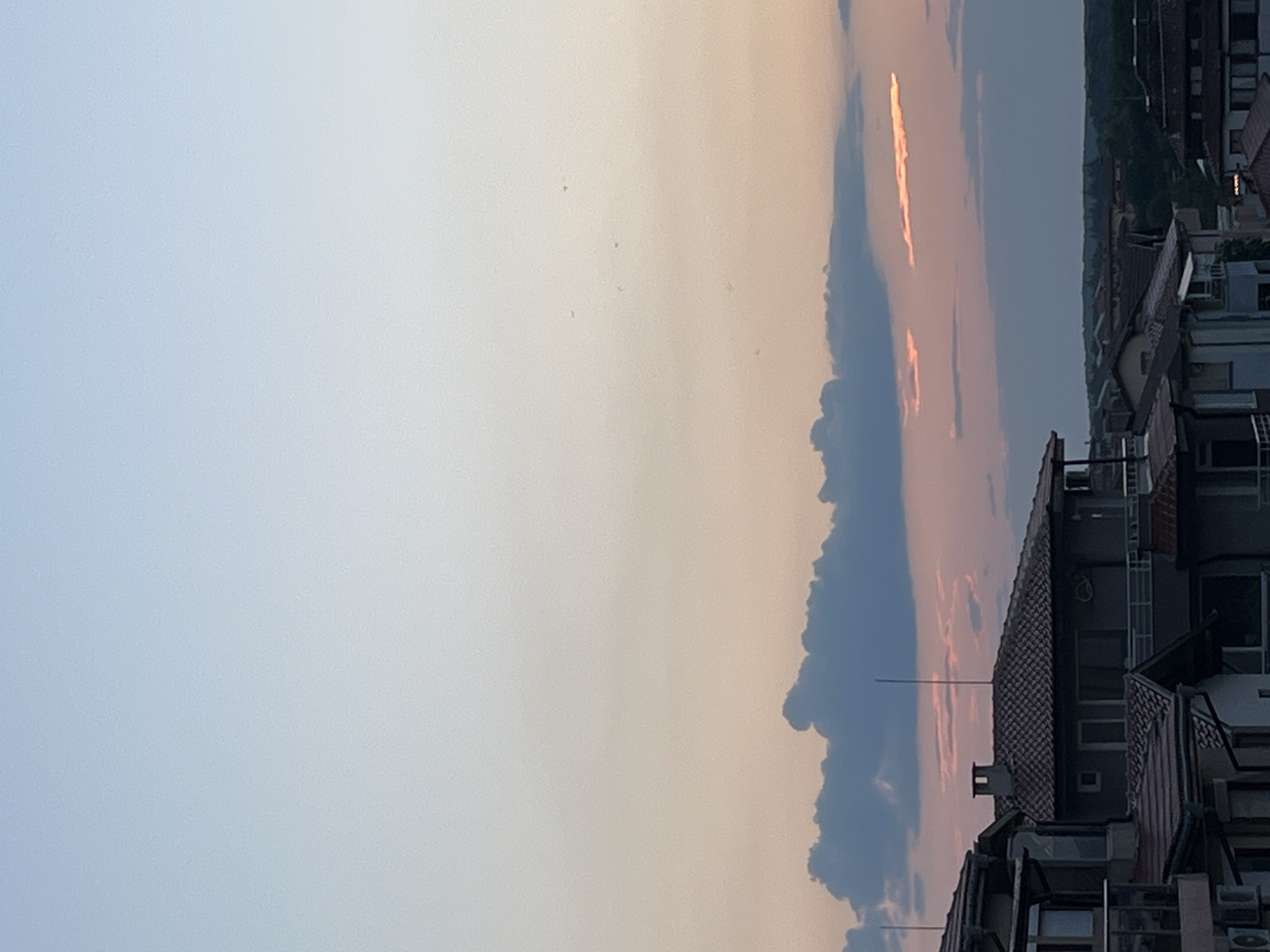 The image shows a sky with a gradient of colors, transitioning from a pale blue at the top to a soft orange and pink hue towards the horizon, indicative of sunset. Several clouds are visible in the sky, some dark and billowy, others wispy and illuminated by the setting sun. Below the sky, the rooftops of several buildings are visible. The architecture appears to be residential, with tiled roofs and balconies. Some of the buildings have prominent chimneys. In the distance, a line of trees can be seen, suggesting a more rural or natural landscape beyond the immediate urban area. The scene is set in the city of Lozenets, Bulgaria. The lighting suggests it is late afternoon or early evening. There are no people or animals visible in the image.