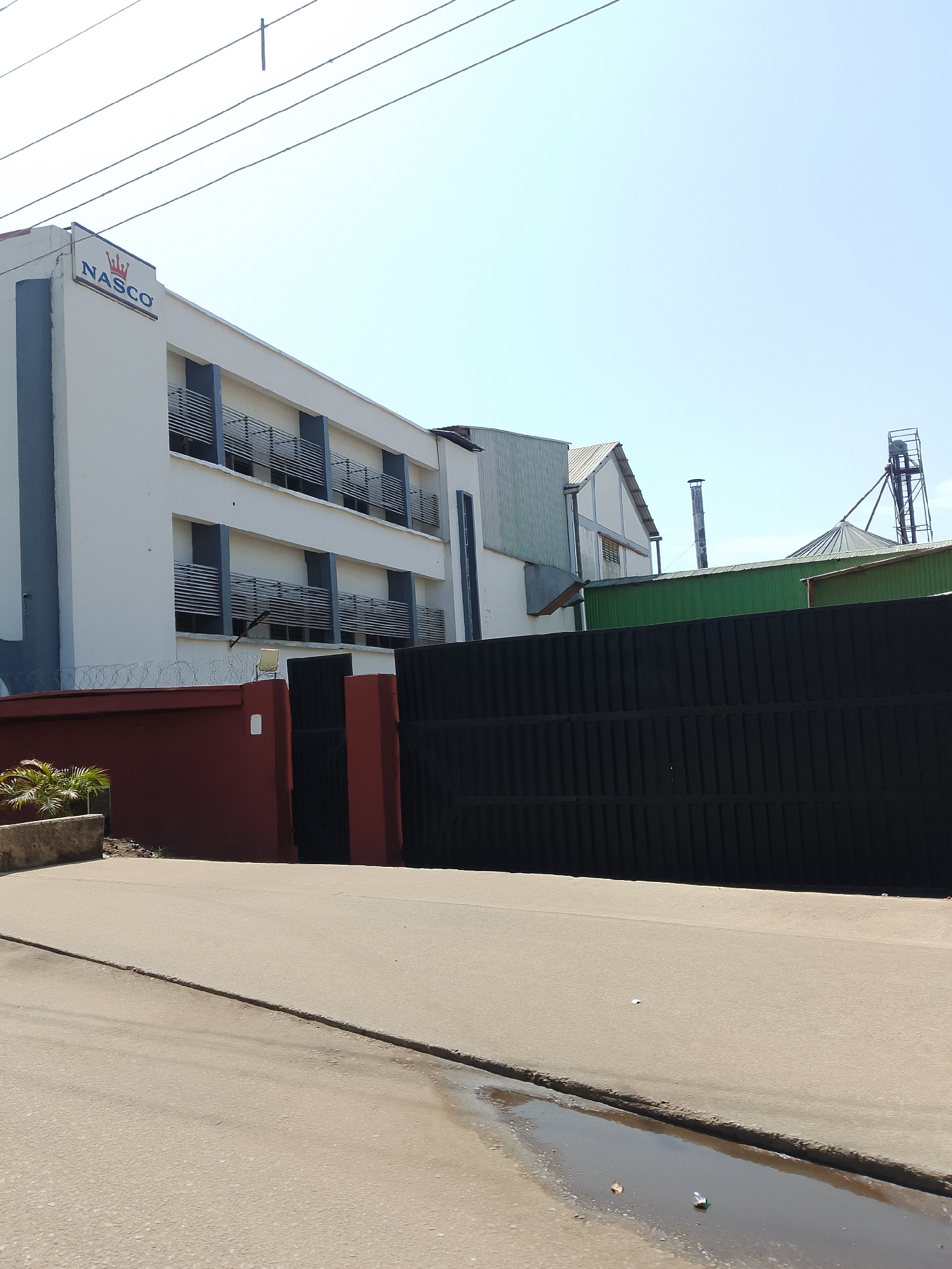 The image depicts a commercial building with a sign that reads "Nasco". The building is white with grey accents and features multiple balconies with horizontal railings. In front of the building is a dark, tall fence, and a red wall. Beyond the main building, other industrial structures are visible, including a silo and a tall metal tower. The scene appears to be in an industrial or commercial area, with a clear blue sky overhead. A paved road runs in front of the property, with a small puddle of water on it. There are no people visible in the image.