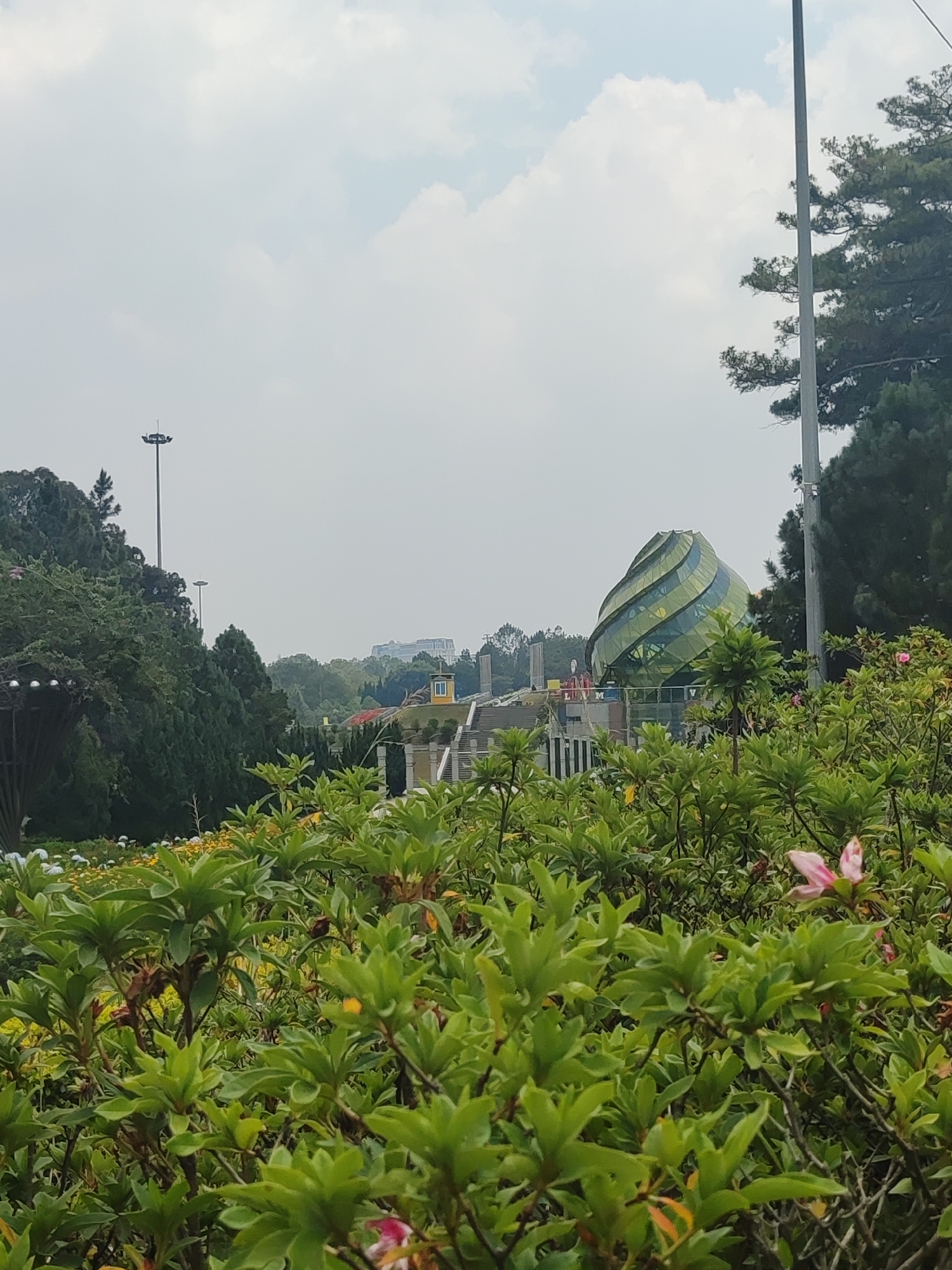 The image shows a partially obscured view of the Artichoke Flower building in Da Lat, Vietnam. The building, characterized by its green, spiraling glass facade, is situated in the background amidst trees and structures, including light poles. The foreground is dominated by lush green bushes with a few visible pink flowers. The sky is overcast with white clouds.