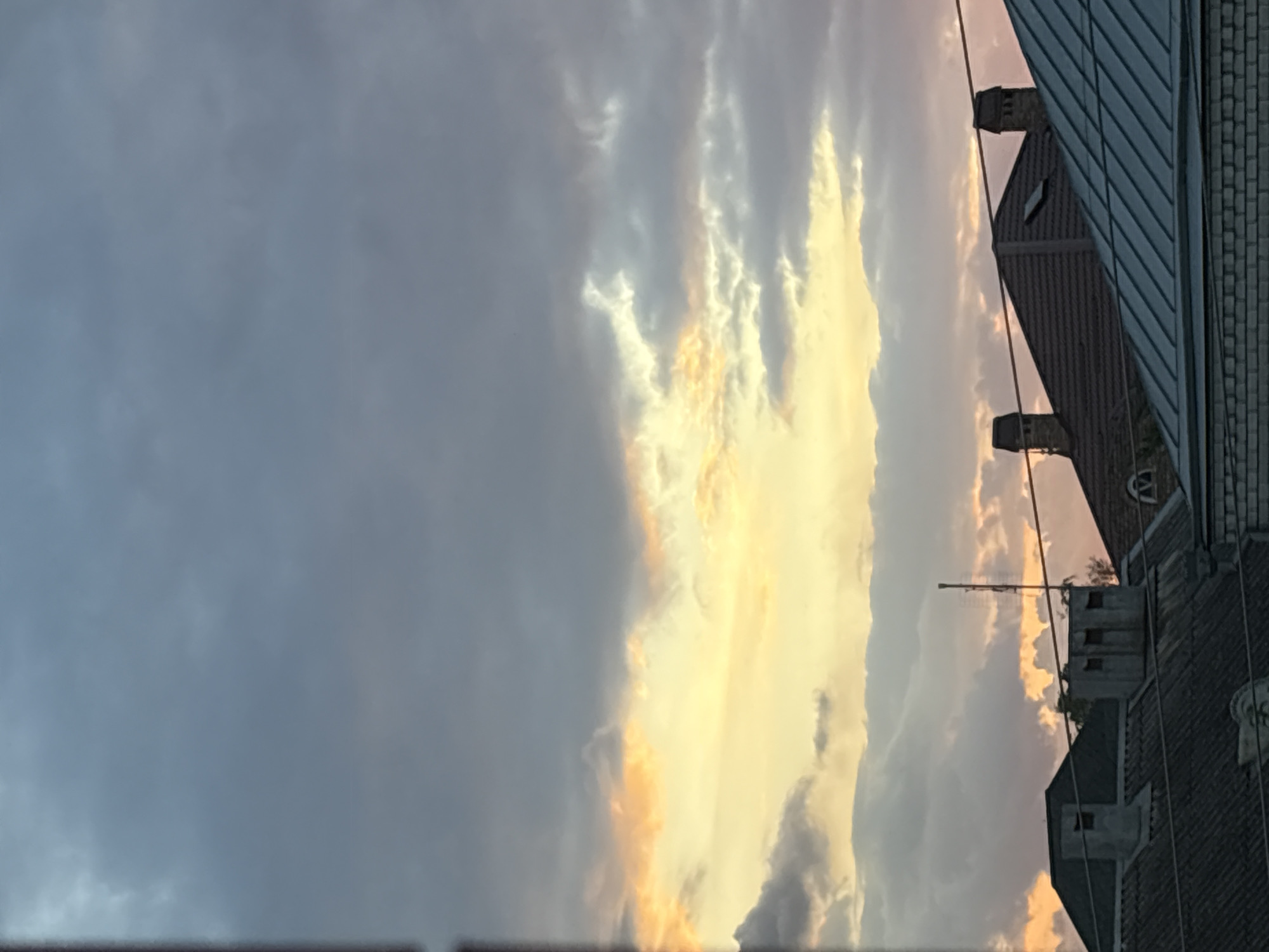 This is a photograph taken from a high vantage point, looking upwards towards a cloudy sky at dusk. The sky is a mix of dark gray and lighter, warmer tones of yellow and pink, indicating the setting sun. Several rooftops with chimneys are visible in the foreground, suggesting an urban or suburban setting. The roofs are made of various materials, including what appears to be dark shingles and corrugated metal. There are also power lines crisscrossing the scene. The overall impression is one of a peaceful, possibly residential, area experiencing the transition from day to night. The specific location is the city of Georgiyevsk, Russia. There are no people visible in the image.