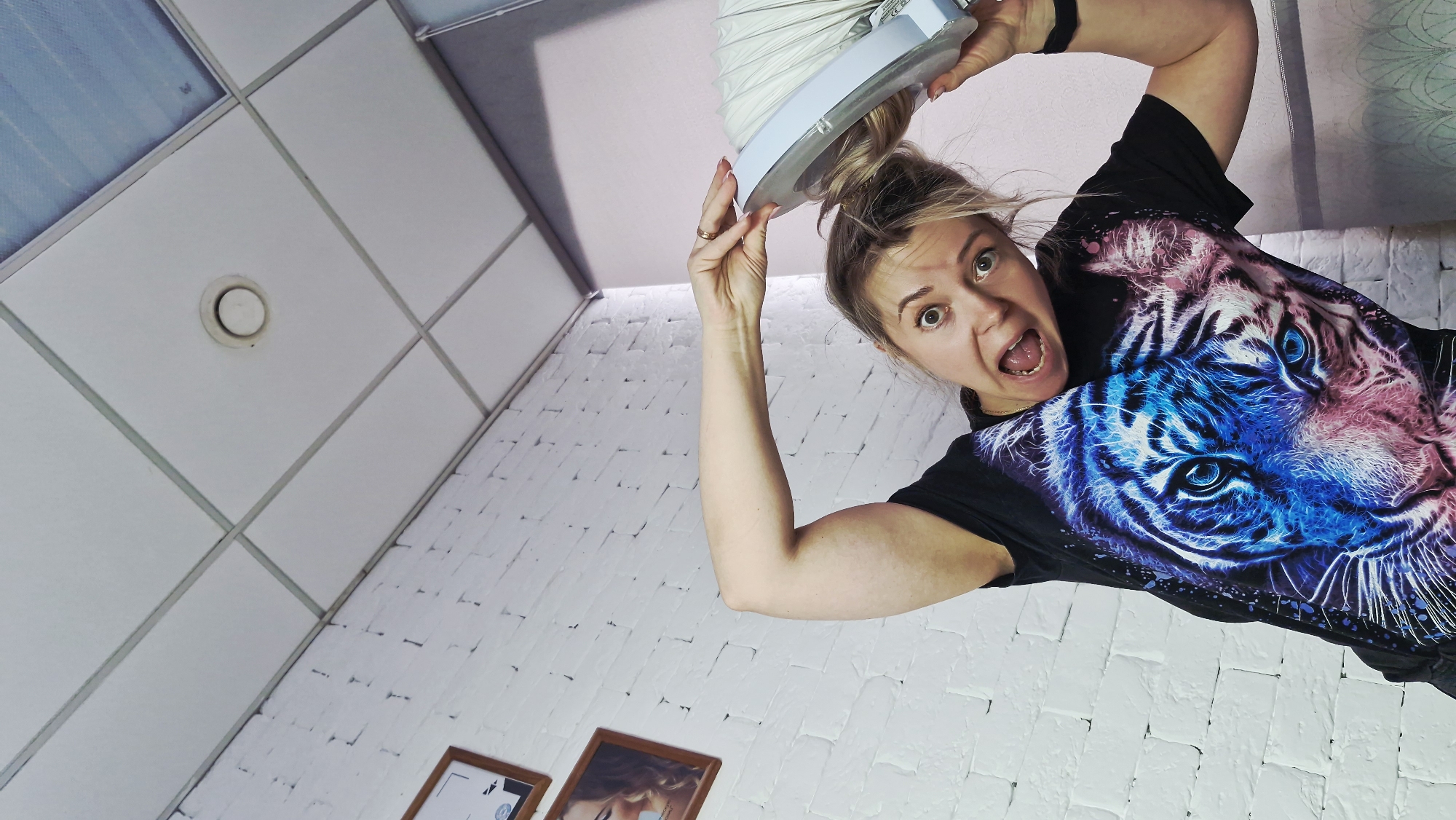 An indoor, low-angle shot captures a young woman looking directly at the camera with wide, surprised eyes and an open mouth, conveying a sense of excitement or playful shock. Her light-colored hair is tied up in a bun, and she wears a black t-shirt featuring a striking, vibrant graphic of a tiger's head rendered in electric blue and pink tones.
She is positioned in the upper right quadrant of the image, appearing to lean into the frame. Her left arm is visible and flexed, while her right hand is raised above her head, holding a white, round, flat object from which a flexible, accordion-style duct extends upwards, mostly obscured by the top edge of the frame. Small, indistinct text, possibly a CE mark, is faintly visible on the white object she holds. She wears a golden ring on her left ring finger and a black wristband on her right wrist.
The background consists of a white drop ceiling with a grid pattern, featuring a small, round white vent or smoke detector in the upper left. Below the ceiling on the right, a white brick-textured wall extends downwards. Partially visible at the bottom edge are two framed pictures hanging on this wall. The unusual upward perspective creates a dynamic and engaging composition, suggesting the camera is placed on the floor or a low surface. The scene takes place indoors in Otchor, Russia.