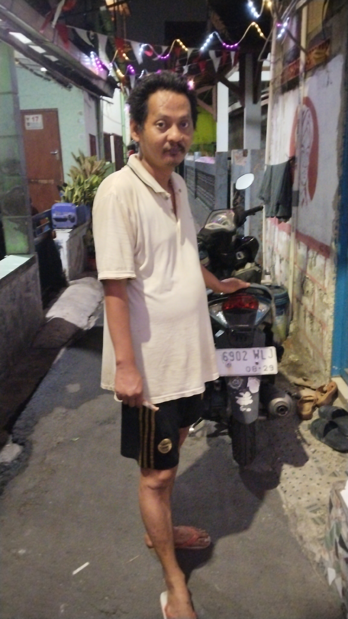 A man stands in front of a motorcycle. He is wearing a light-colored short-sleeved polo shirt and black shorts with yellow stripes. The motorcycle is parked on a paved surface, possibly an alley or street, in front of a building. The building has a textured wall with some debris and laundry hanging on it. There are decorative lights strung overhead. The man is holding a cigarette in his right hand. The license plate on the motorcycle reads "6902 WLJ" and "08-29". The overall scene suggests a residential area in a city in Indonesia, likely South Tangerang given the presence of Indonesian script on a sign in the background, though the text is too blurry to read definitively.
