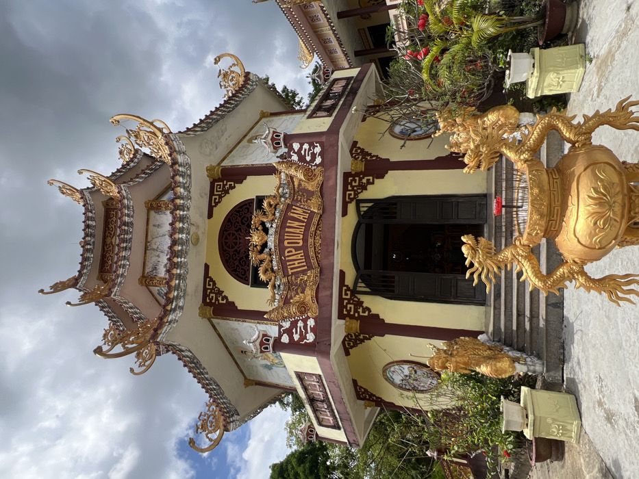 This image depicts the Tháp Quan Âm (Quan Am Pagoda/Tower) located in Đăk Mar, Vietnam. The central subject is a multi-tiered, pagoda-style building with pale yellow walls, dark red trim, and extensive golden ornamentation.
The structure features a complex, tiered roof with upward-curving eaves adorned with intricate golden designs and finials. Below the main roofline, the upper level displays decorative panels, some appearing to be murals or relief sculptures, bordered by white and red elements. The second level includes a balcony-like section with a dark red railing.
The main entrance on the ground level consists of dark double doors, slightly ajar, revealing a dim interior. Above the entrance, a prominent golden sign with the Vietnamese text "THÁP QUAN ÂM" is flanked by ornate golden dragon sculptures. A large, intricate arched window or decorative panel is situated above this sign.
In the immediate foreground, a substantial golden incense burner (urn) is centrally positioned. It is adorned with two golden dragon figures serving as handles or supports and contains numerous thin, dark incense sticks with a single red element, possibly a candle or larger incense stick, protruding.
Stone steps, flanked by golden dragon-shaped railings, lead up to the pagoda's entrance. Surrounding the base of the building are various potted plants, some with green foliage and others displaying red flowers. A light-colored, pedestal-like planter sits to the left of the steps.
The ground in front of the pagoda is a clear, light-colored paved surface. Green trees are visible on the left and right sides of the building, suggesting a vegetated setting. A partially visible structure with a similar architectural style is present on the far right. The sky overhead is partly cloudy. No individuals are present in the frame.