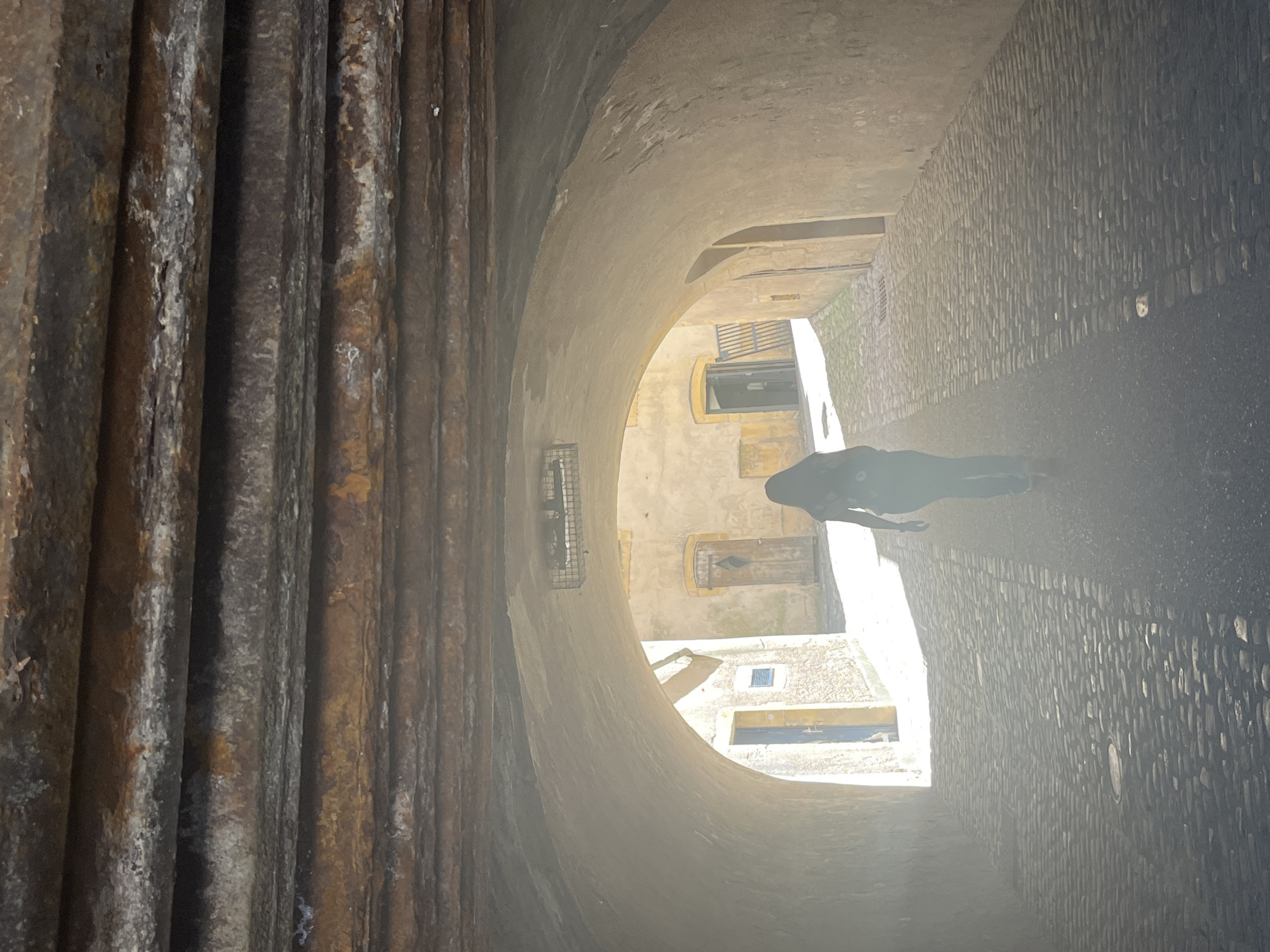 The image depicts a person walking through an arched stone passageway in Metz, France. The passageway is lined with textured walls and a stone ceiling. The floor is paved. The light at the far end of the passageway is bright. Details of buildings are visible through the archway. The person's form is somewhat indistinct due to the bright light. The scene seems to be in the old part of the city.