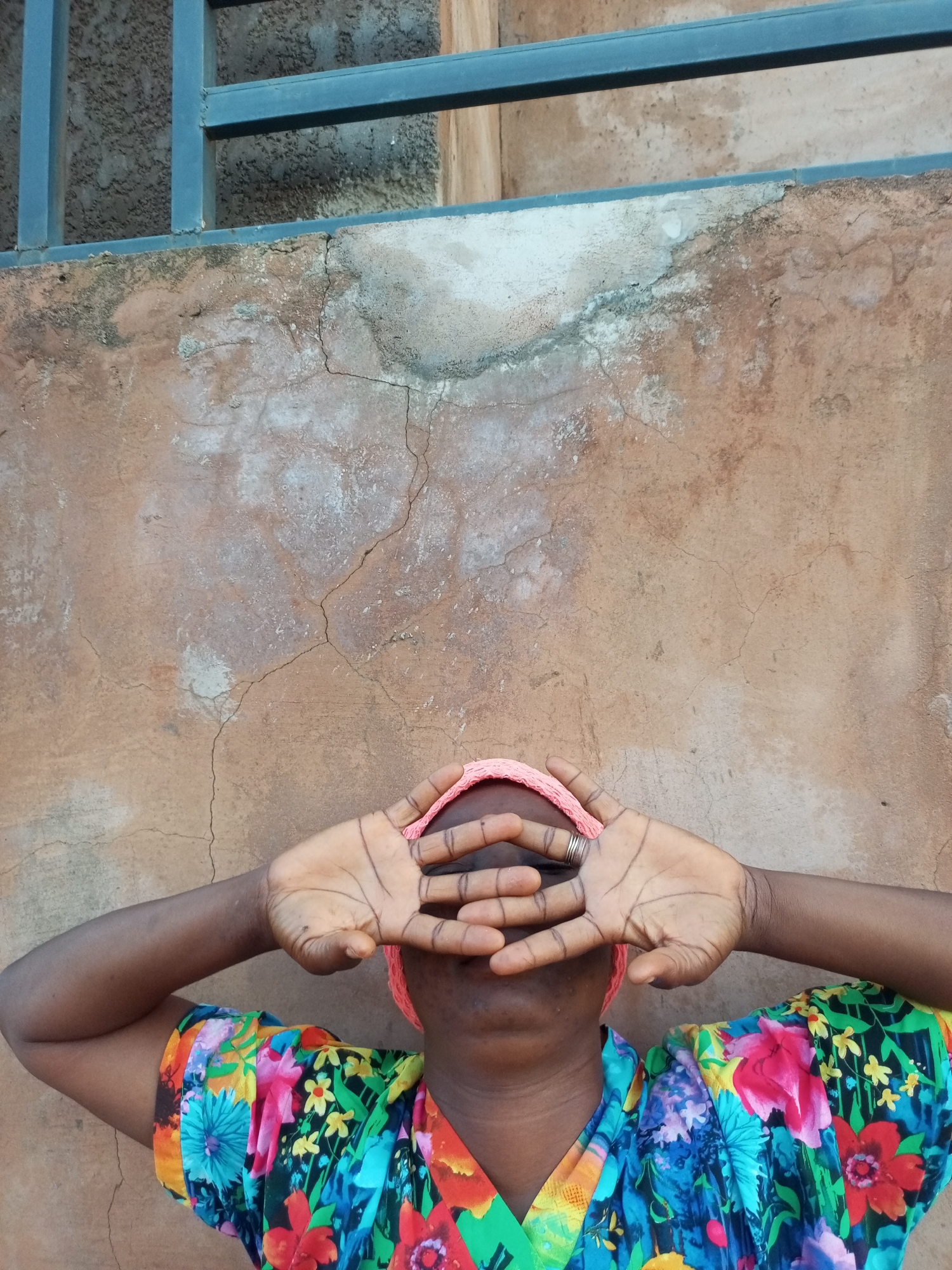 A close-up, head-on, eye-level shot shows a person's upper body against a textured, tan-colored wall. The person, who has dark skin, is wearing a brightly colored, floral-print shirt with a V-neck. They are also wearing a pink headscarf, and their hands are held up to cover their eyes, with fingers spread. A ring is visible on the middle finger of their right hand. Above the wall, a dark grey metal railing runs horizontally. The wall shows signs of age, with visible cracks and patches of lighter discoloration. The scene appears to be outdoors, possibly in a residential or public area in Tempure, Nigeria.