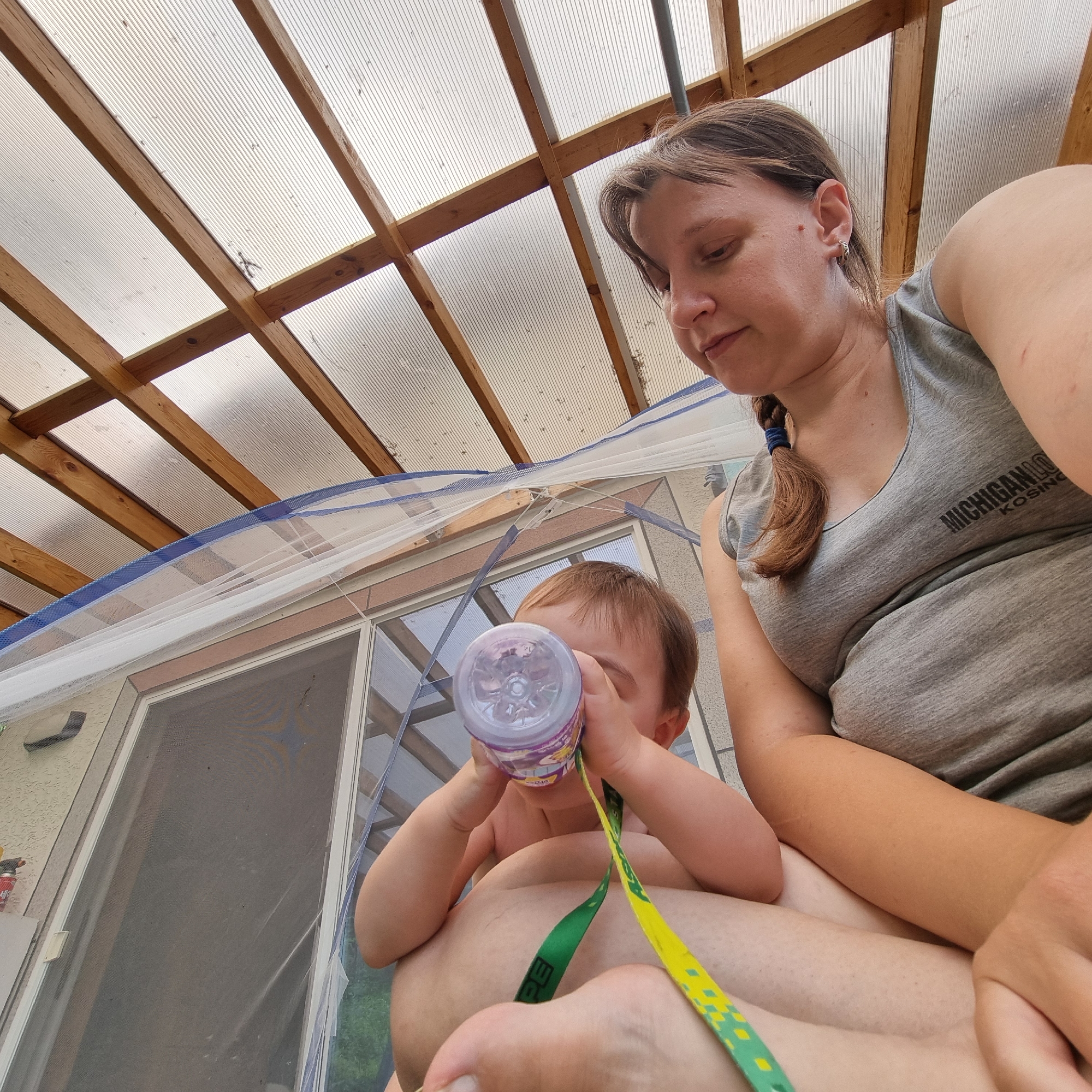 The image captures a mother and child in an outdoor, covered seating area, possibly a sunroom or porch, with a corrugated plastic roof supported by wooden beams. The mother, wearing a gray t-shirt with "MICHIGAN LO KOSINO" printed on it, is seated, with her arm around the child. The child, facing away from the camera and towards a bottle, appears to be drinking from it. The child is wearing a green and yellow ribbon, possibly attached to the bottle. The background shows a glass door with a screen and some vegetation outdoors. The overall lighting suggests it is daytime. The image does not contain any information that would allow for pinpointing the location to Dunnae-myeon, South Korea.