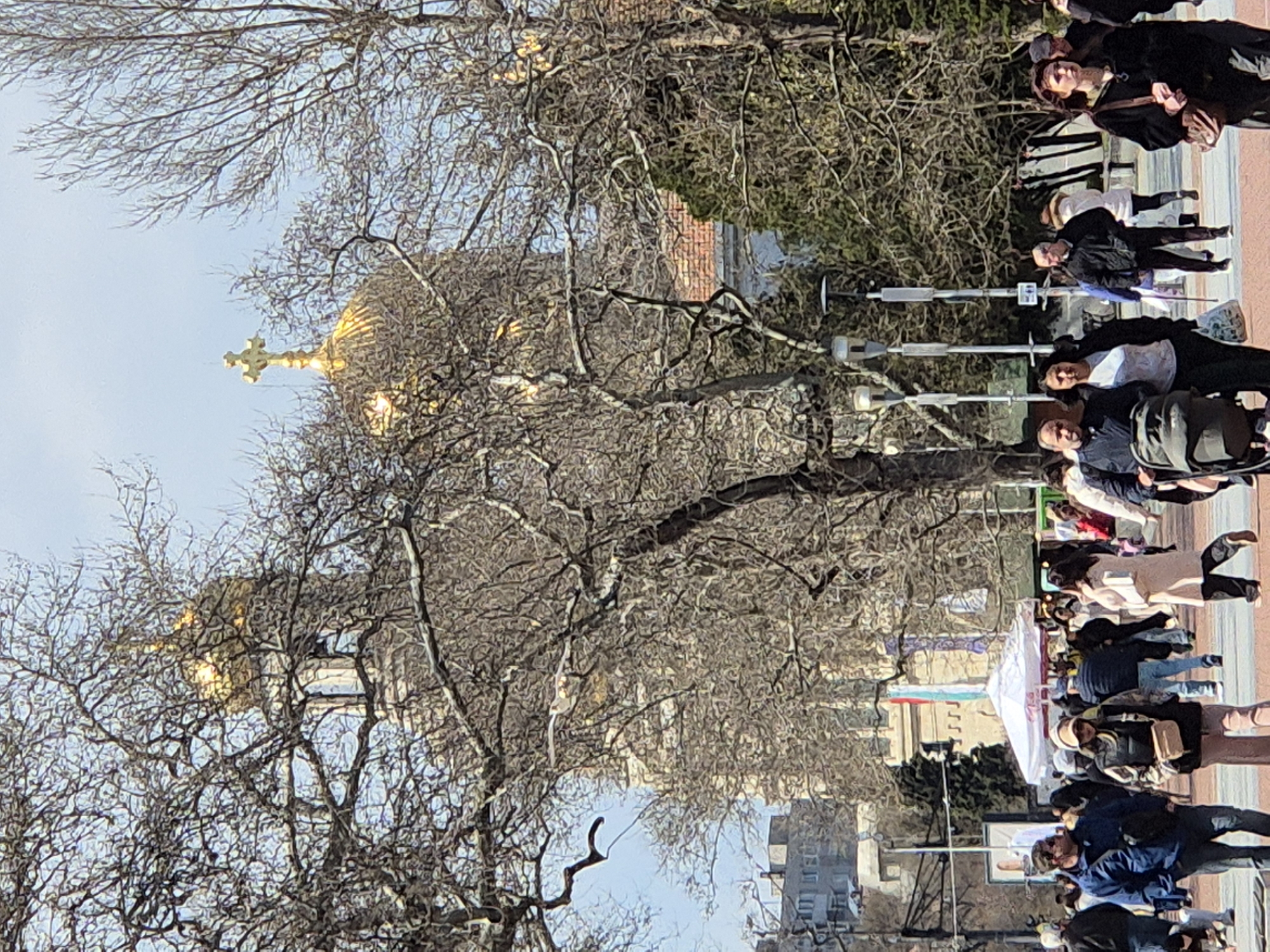The image shows a street scene in an urban location, likely in Unknown, Unknown. In the foreground, a bare tree fills much of the left side of the frame. In the background, there is a large building with a prominent golden dome and a cross on top. People are walking along the right side of the image. There are also streetlights and other buildings visible in the background. The sky is overcast.