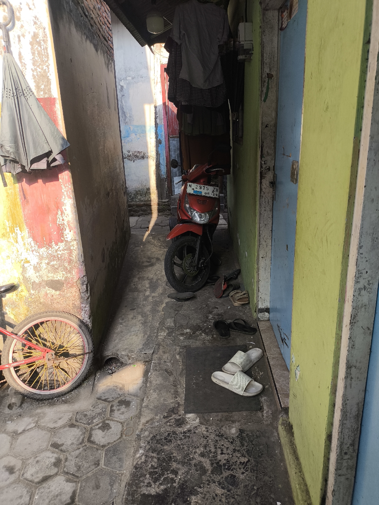 The image depicts a narrow alleyway in the city of Citrodiwangsan, Indonesia. The alley is bordered by two buildings, one with a rough, red and yellow painted wall on the left, and another with a brightly painted green and blue door on the right. Sunlight streams down the alley, casting strong shadows.

On the left side, a folded dark umbrella is propped against the red and yellow wall. A bicycle with a red frame and yellow spokes is positioned in the foreground, its wheel visible. The ground of the alley is paved with hexagonal stones and patches of concrete.

In the center of the alley, a red motorcycle with a license plate reading "N 2975 ZH 05.29" is parked. In front of the motorcycle, several pairs of footwear are scattered on the ground, including black flip-flops and light-colored slide sandals. The wall of the building on the right has peeling paint and what appears to be an electrical box mounted on it. Clothing is hanging on a line above the motorcycle. The overall scene suggests a lived-in, utilitarian space within a residential area.