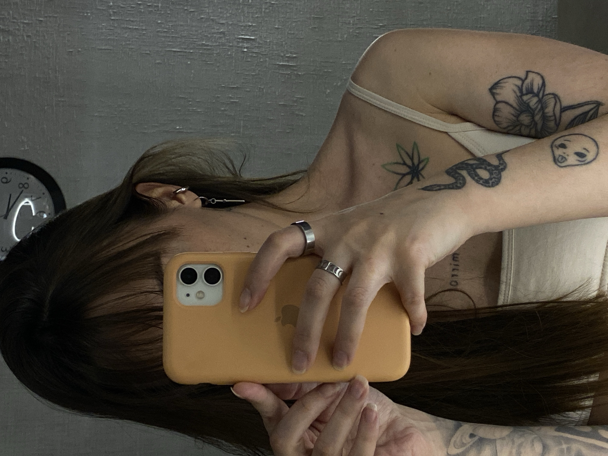 A person is holding an orange-cased iPhone in a selfie pose. The person has dark hair with bangs obscuring part of their face. Multiple tattoos are visible on their arms and shoulders: a flower, a marijuana leaf, a snake, and a small face. The person is wearing silver rings. In the background, a clock hangs on a wall, and the wall appears to have a textured or peeling surface. There is no information available from the image to indicate that the image was taken in Votkinsk, Russia.