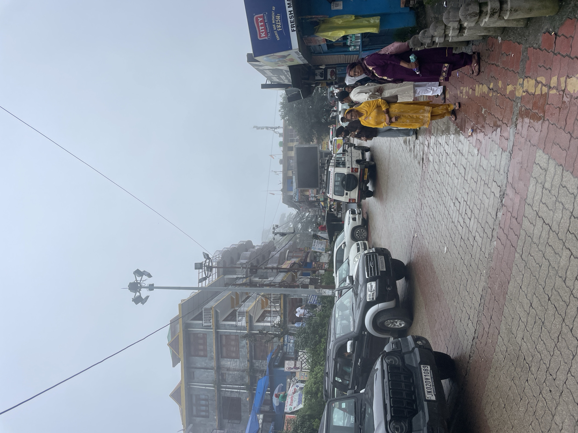 This is a street scene in Bhagsu, India, on a foggy or overcast day. The street is paved with red and gray interlocking bricks, which appear wet, indicating recent rain. Several vehicles are parked and driving on the street, including a black Mahindra Thar in the foreground on the left, a black Ford Endeavour in the center-left, and a white Bolero SUV in the background. Buildings with multiple stories line the right side of the street, featuring balconies and awnings. Some of these buildings house shops, including a bakery called "Kitty The Master Baker" with a blue facade. On the left side of the street, there are more buildings, some with blue awnings and posters displayed. A tall lamppost with multiple lights is visible near the center of the image. A group of people are standing on the right side of the street near the shops, including a woman in a yellow outfit, a man in a white kurta, and a woman in a purple outfit. Another person holding an umbrella is visible further down the street under a blue awning. The overall atmosphere is somewhat hazy due to the weather conditions.