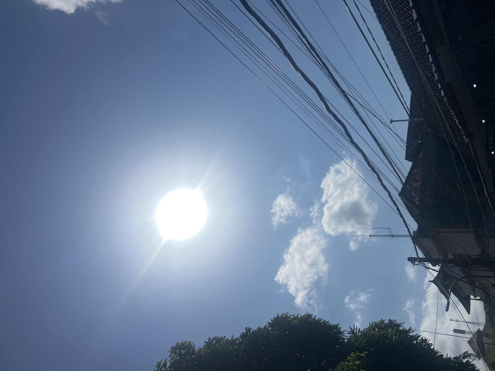 In Kuta, Indonesia, on a bright, sunny day, the clear blue sky is dominated by a brilliant sun, casting a strong glare. A few scattered white clouds drift by. In the foreground, a lush green tree occupies the left side of the frame, its leaves dense and verdant. The scene also features several buildings with traditional tiled roofs, some showing signs of age. A tangle of electrical wires crisscrosses the upper right portion of the image, running diagonally against the sky. Various antennas and poles are visible atop the buildings, suggesting a populated area. The overall impression is one of a typical urban streetscape under intense tropical sunlight.