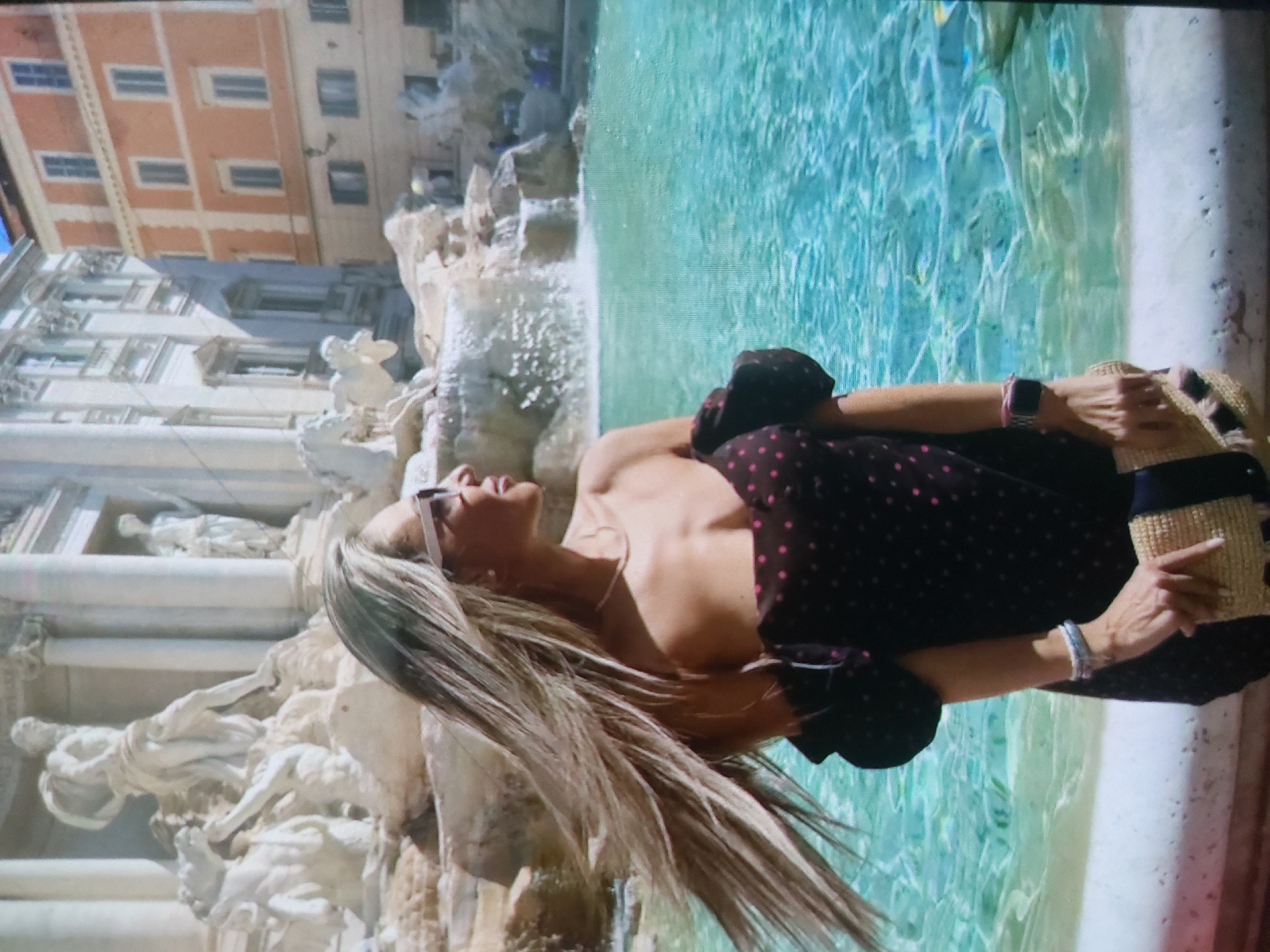 A woman with long, blonde hair, wearing sunglasses, a black and pink polka dot dress, and a smartwatch, is lying on the edge of the Trevi Fountain in Rome. She is holding a straw hat. The fountain has sculptures and clear, blue water. Buildings are visible in the background.