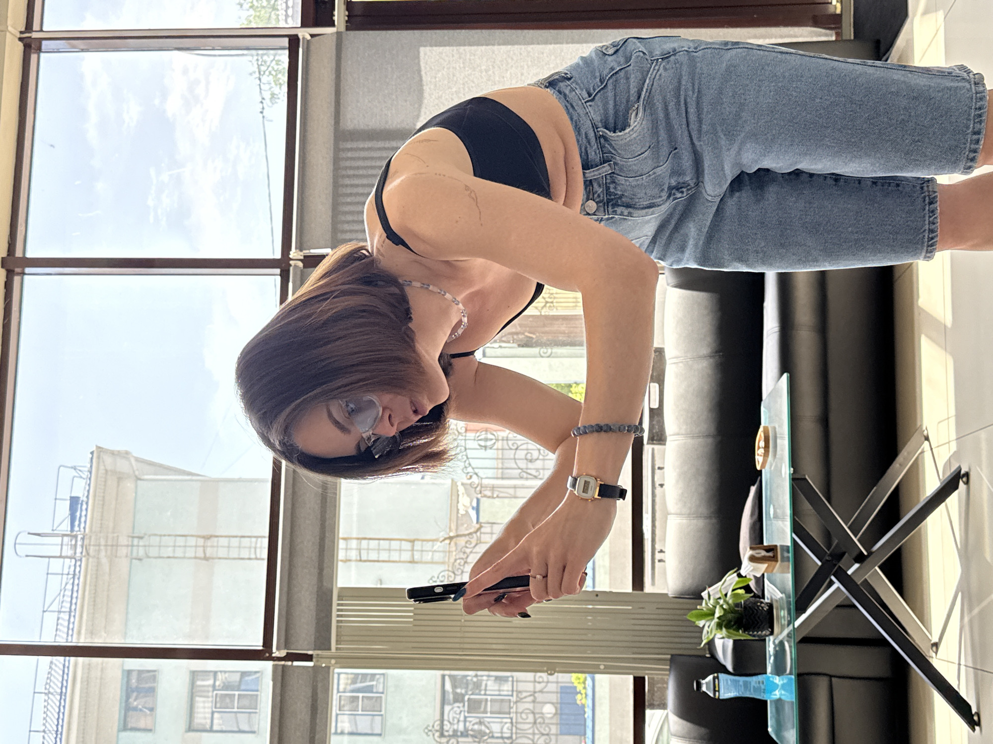 A woman in Rostov-on-Don, Russia is bending over while holding a smartphone. She is wearing sunglasses, a black top, and denim shorts. She has short brown hair. She is wearing a bracelet and a watch on one wrist, and a necklace. She is standing near a glass table with a plant, a decorative object, and a bottle of blue liquid. Behind her is a black leather couch. Large windows show a view of buildings and the sky. The building visible outside the window is approximately two stories tall.