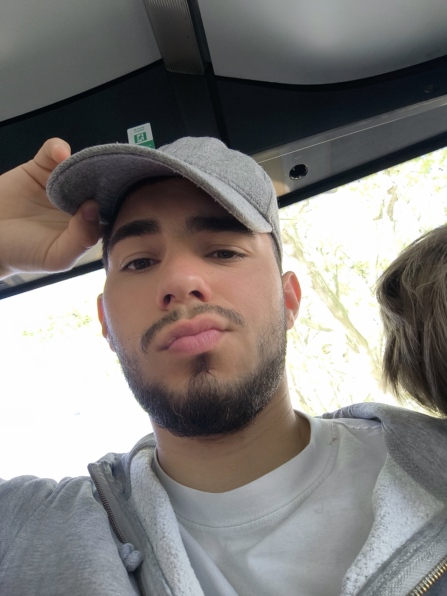 A close-up selfie shows a young man inside what appears to be a public transport vehicle, likely a bus, during daylight hours. He is centered in the frame, looking directly at the camera with a neutral to slightly pouting expression. He has dark hair, dark eyes, and a neatly trimmed beard and mustache.
He is wearing a light grey baseball cap, which he is holding with his left hand, a grey hooded sweatshirt with a zipper, and a white crew-neck t-shirt. The bright sunlight from outside illuminates his face.
Through the vehicle's window, visible on the right side and upper left, are blurred green trees, indicating an outdoor, possibly park-like, environment and a sunny day. Part of another person's head with brown hair is visible on the far right edge of the image.
Above the man's head, the interior of the vehicle is visible, featuring a grey ceiling and a panel above the window. A green emergency exit sign with the universal "running man" symbol is distinctly visible on an interior panel on the left, slightly obscured by the cap. The setting is consistent with public transportation in Strasbourg, France. No other discernible text is visible.