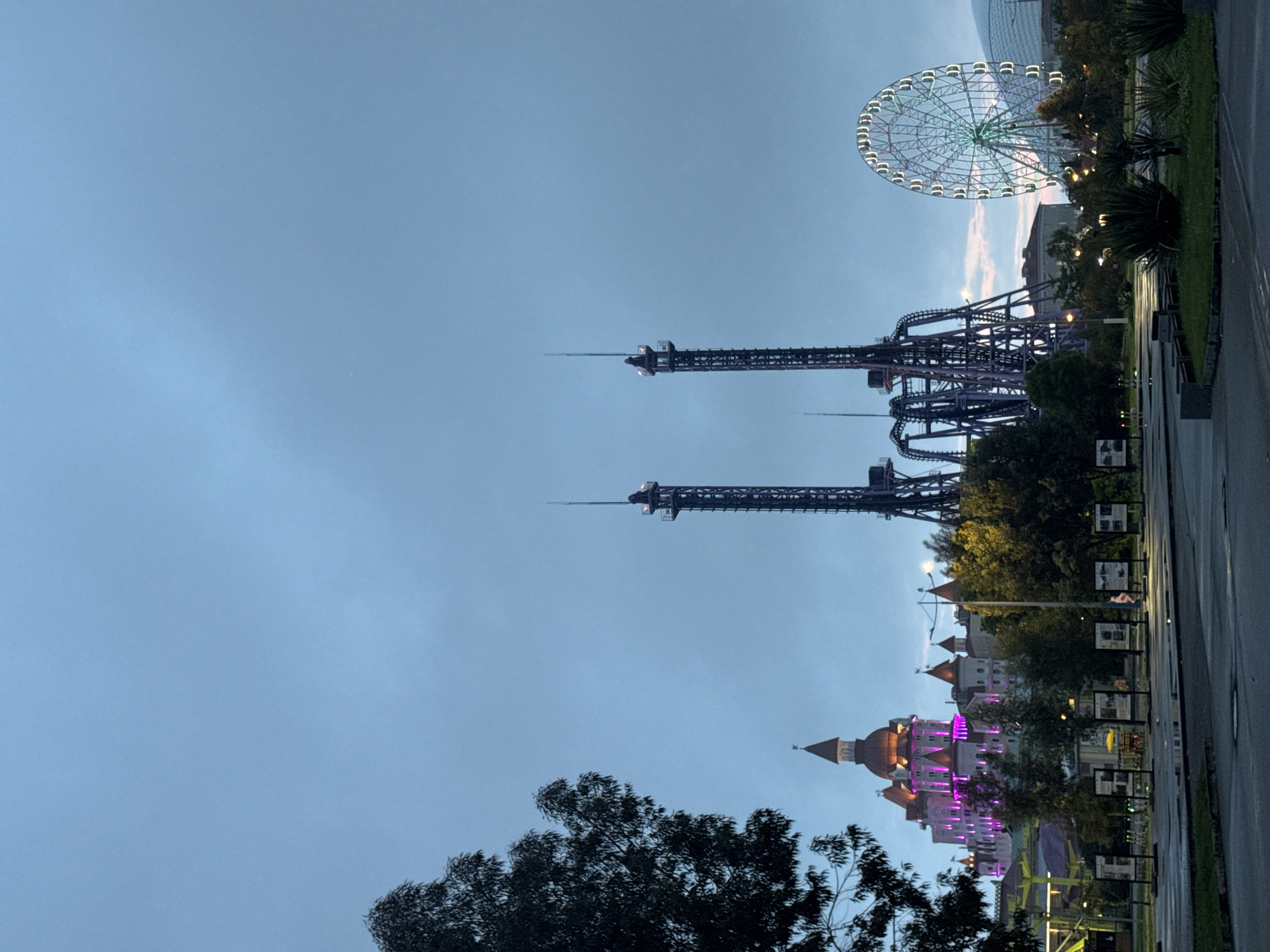 The image shows a view of Sochi Park in the city of Sirius, Russia. The park features several amusement park rides, including a large Ferris wheel, two drop towers, and a roller coaster. In the foreground is a building with multiple towers and pink lighting. Trees are partially obscuring the view of the building. The sky is overcast.