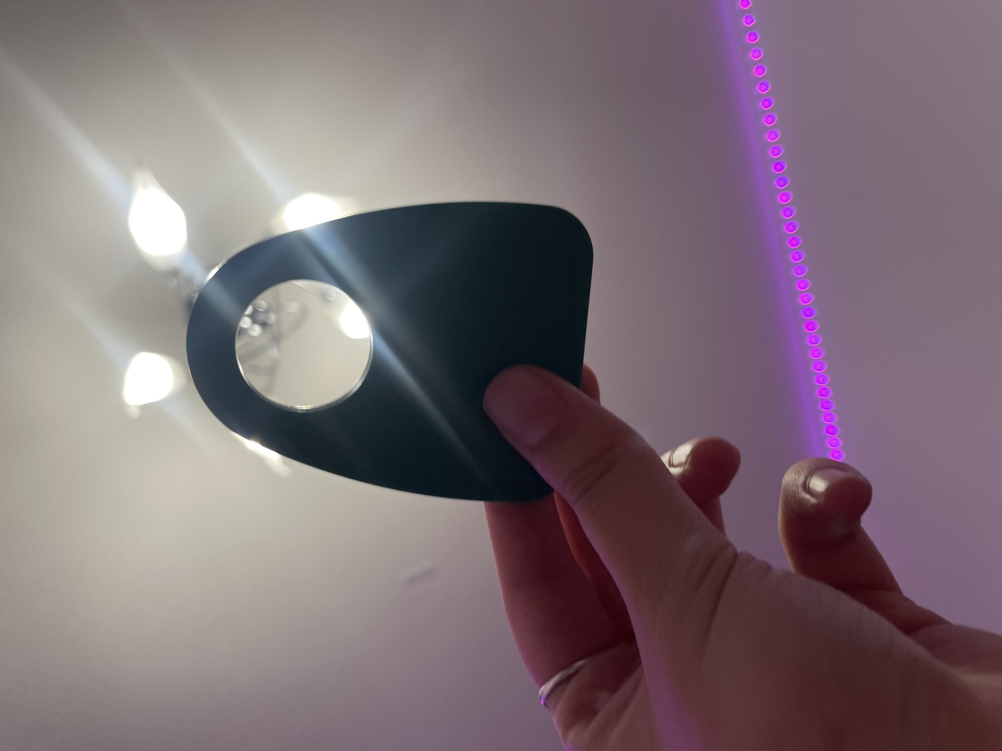 A hand, wearing a silver ring on the index finger, holds a dark, tear-drop-shaped object with a circular cutout. The object is positioned against a ceiling with visible light fixtures emitting bright light. Below the hand, a string of pink LED lights is visible, creating a gradient of pink and purple light. The background appears to be an indoor setting. There is no indication of the location being Vaganovo, Russia.