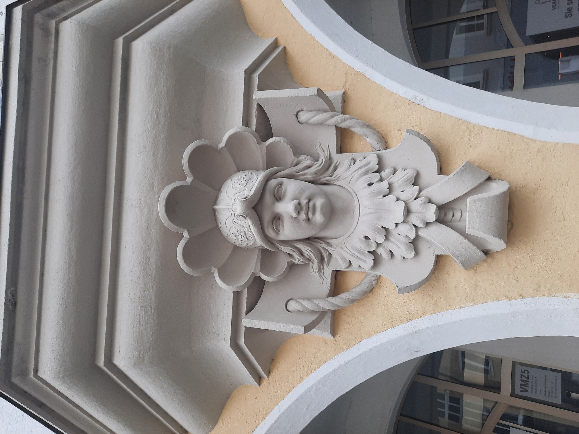 The image shows an exterior architectural detail on a building in Rosenheim, Germany. The focal point is a bas-relief sculpture of a person's head and shoulders, adorned with a helmet and decorative elements. The sculpture is rendered in a light-colored material, possibly stone or stucco, and is mounted on a textured, yellow-colored wall. Above the sculpture is an ornate, layered architectural element. To the left and right of the sculpture, portions of arched doorways or windows are visible, with glass panes reflecting interior spaces. A sign with German text, including "VMZ5," is affixed to the window on the left. The overall impression is of a historical or ornate building facade.