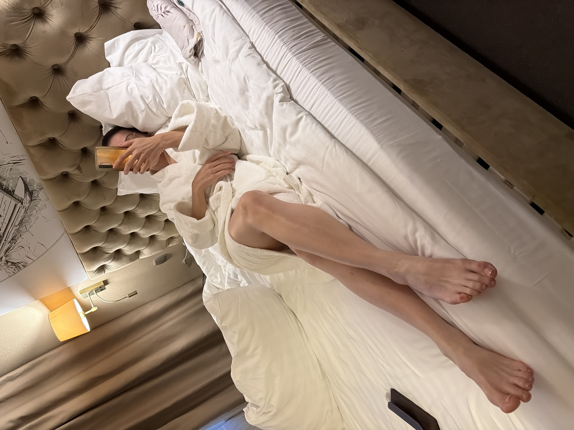 A high-angle, slightly skewed photograph captures a person lying on a bed, predominantly covered by a white duvet and wearing a white bathrobe. The person is using a light-colored smartphone, held up to their face, partially obscuring it. Their left hand holds the phone, while their right hand rests on their chest. The person's bare legs are visible extending out from the bathrobe and duvet, with their feet crossed at the ankles.
The bed features a tufted, light-brown headboard on the left side of the frame, and a minimalist bed frame or side table is visible on the right. Below the headboard, a white wall is adorned with a black and white artistic print. A bedside lamp with a warm-toned shade is mounted on the wall below the print, casting a soft glow. The room appears to be indoors, likely a bedroom or hotel room, given the plush bedding and style. The lighting suggests it might be evening or nighttime. No specific emotions are discernible due to the person's face being partially covered. There is no visible text in the image.
