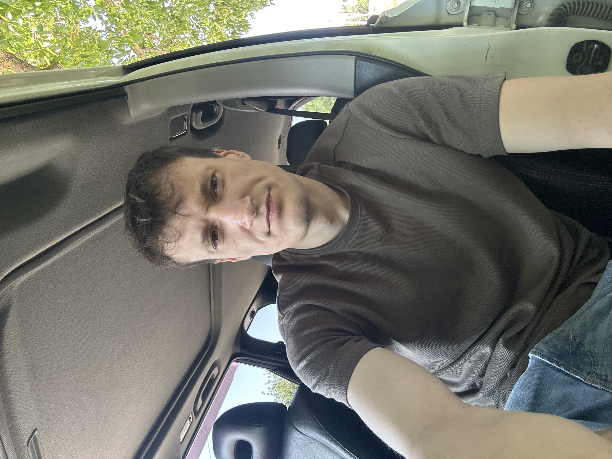 A young man with short brown hair and blue eyes is taking a selfie inside a car. He is wearing a brown t-shirt and blue jeans. The car's sunroof is open, and the sky is visible through it. Green trees and foliage can be seen through the open car door and the windows. The image appears to be taken outdoors on a sunny day, possibly in Makiv, Ukraine, given the context provided. The interior of the car is visible, including the seats and the ceiling.