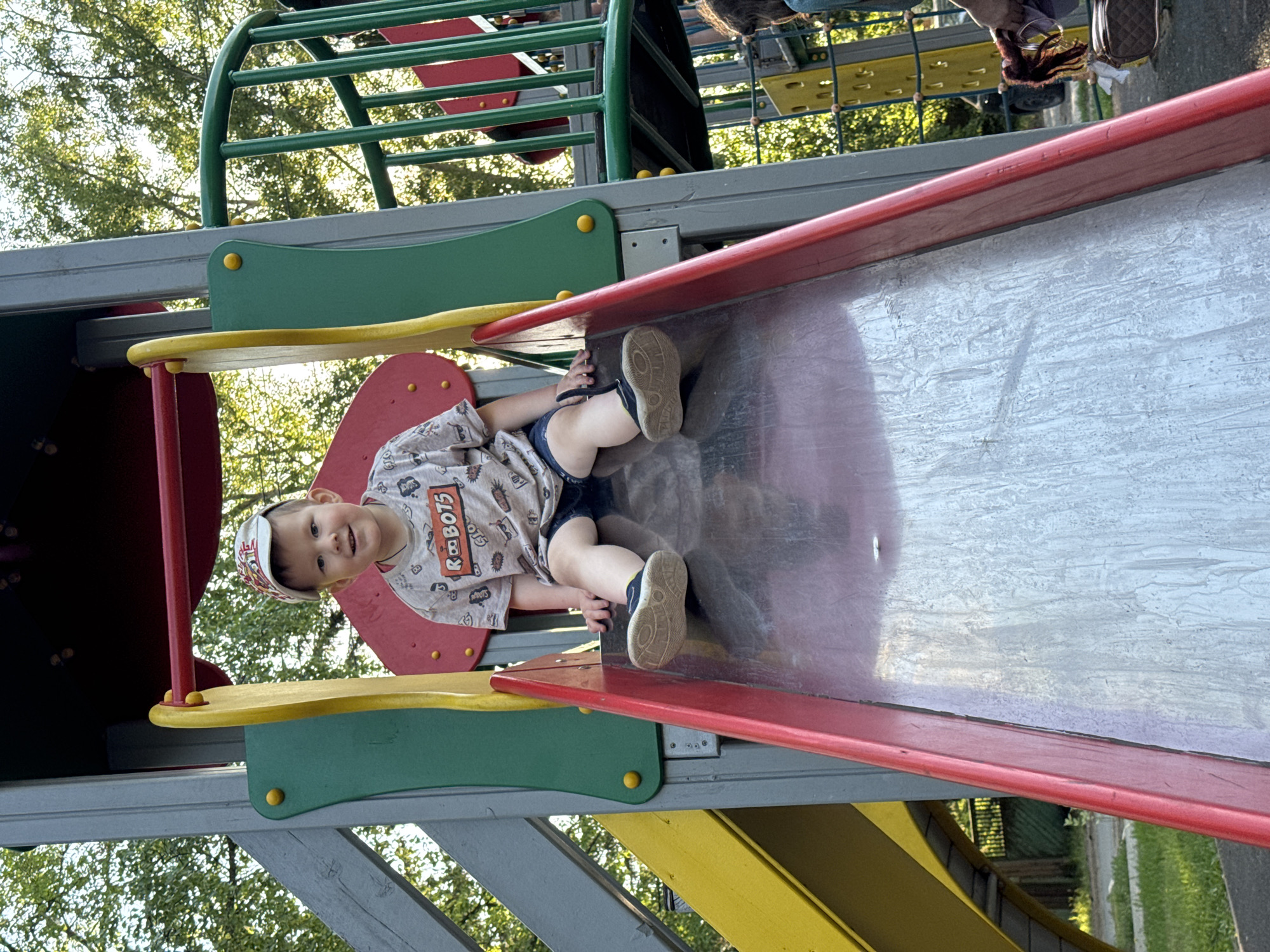 A young child is seated on a playground slide. The slide is constructed with red and gray railings and a reflective metal surface. The child is wearing a light-colored shirt with a "ROBOTS" graphic, dark blue shorts, and a patterned baseball cap. The playground equipment includes green, yellow, and gray support structures. In the background, trees and a glimpse of other playground features are visible. The location is likely a playground in Zav'yalovo, Russia.