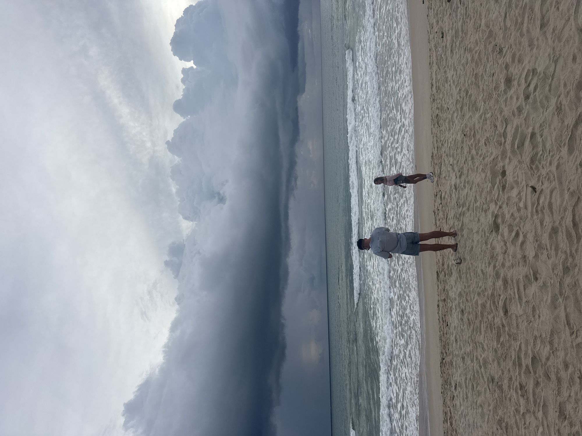 The image shows a wide, sandy beach in Choeng Thale, Thailand, with two individuals standing near the water's edge. The sky is filled with dramatic, dark, and turbulent storm clouds, suggesting an impending downpour. The ocean is choppy with significant waves breaking on the shore.
In the foreground, a person, possibly a man, is standing with their back to the camera, wearing a light-colored short-sleeved shirt and denim shorts. They appear to be holding a phone to their ear. To the right of this person, a young woman, seen from the side, is wearing a pink long-sleeved top and shorts, facing the water. A small, light-colored object, possibly a ball or a shell, is on the sand near the man's feet. The sand itself is light brown and textured, with visible ripples.
The ocean is a mix of deep green and grey, reflecting the overcast sky. The waves are white-capped and energetic. In the distance, a visible streak of rain falls from the dark clouds over the sea, creating a hazy effect on the horizon. The overall atmosphere of the image is moody and dramatic due to the stormy weather.
