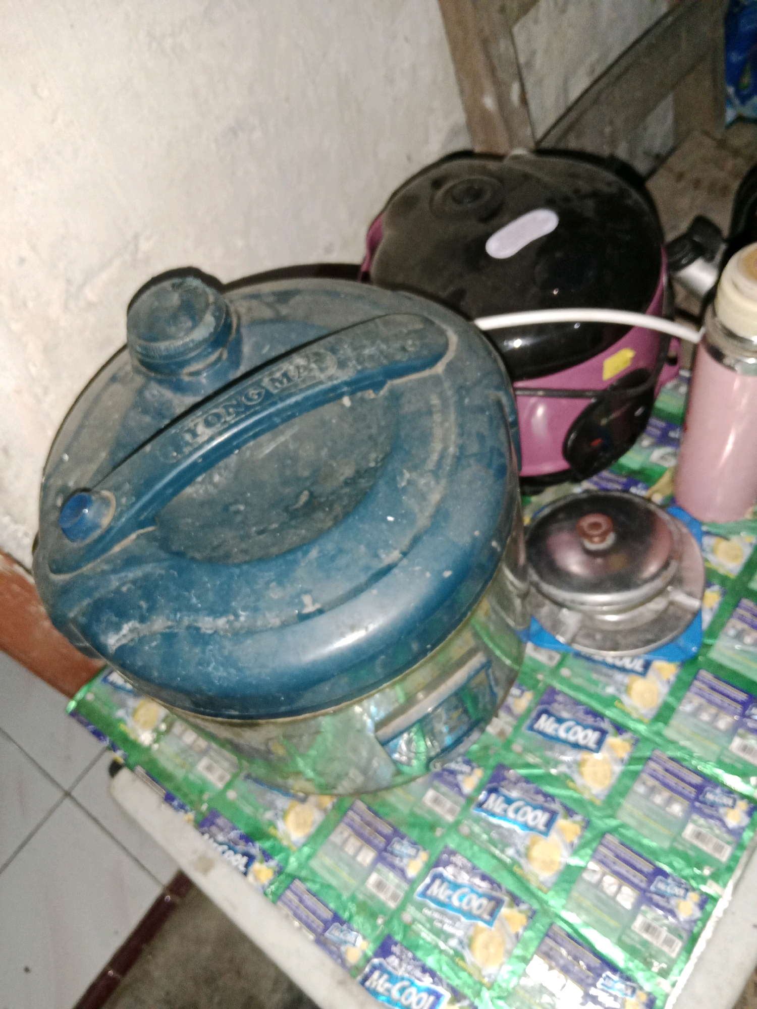 This is a forensic, matter-of-fact description of the image. The image depicts a collection of household objects on a table in what appears to be a residential setting in Ringinrejo, Indonesia.
In the foreground, a large, dusty, blue plastic container with a handle is prominently displayed. The container has a cap and a spout. To the right of this container, a small, metallic object, possibly a lid or a component from another appliance, sits on a surface covered with individually wrapped items. These items are green and feature the "McCOOL" logo, suggesting they are food products, likely snacks or beverages.
Behind the blue container, a black and pink electric appliance, possibly a rice cooker, is visible. It has a rounded lid with a light-colored marking. To the far right, a pink cylindrical object, potentially a thermos or a bottle, stands upright. The background shows a textured wall and wooden structures, indicating an indoor environment. The overall scene suggests a storage or preparation area within a home. No individuals are present in the image.