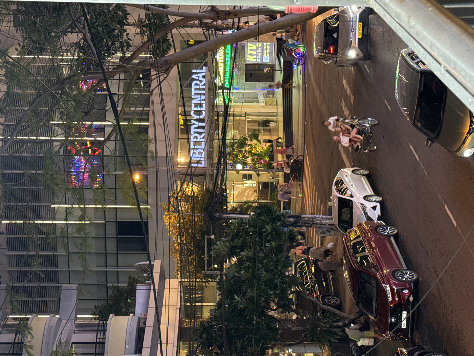 The image captures a nighttime street scene in front of the Liberty Central Nha Trang hotel in Nha Trang, Vietnam. The hotel building is prominently lit with vertical white lights and illuminated signage. In the foreground, a maroon SUV is parked on the right side of the road, facing left. To its left, a white sedan is parked. Further down the street, a silver sedan is in motion. A three-wheeled vehicle, possibly a cyclo or tuk-tuk, with people on board is also visible on the road. Pedestrians are seen walking on the sidewalk and crossing the street. The environment suggests a busy urban area with a mix of vehicles and people. Trees and overhead power lines are also present, contributing to the city atmosphere.