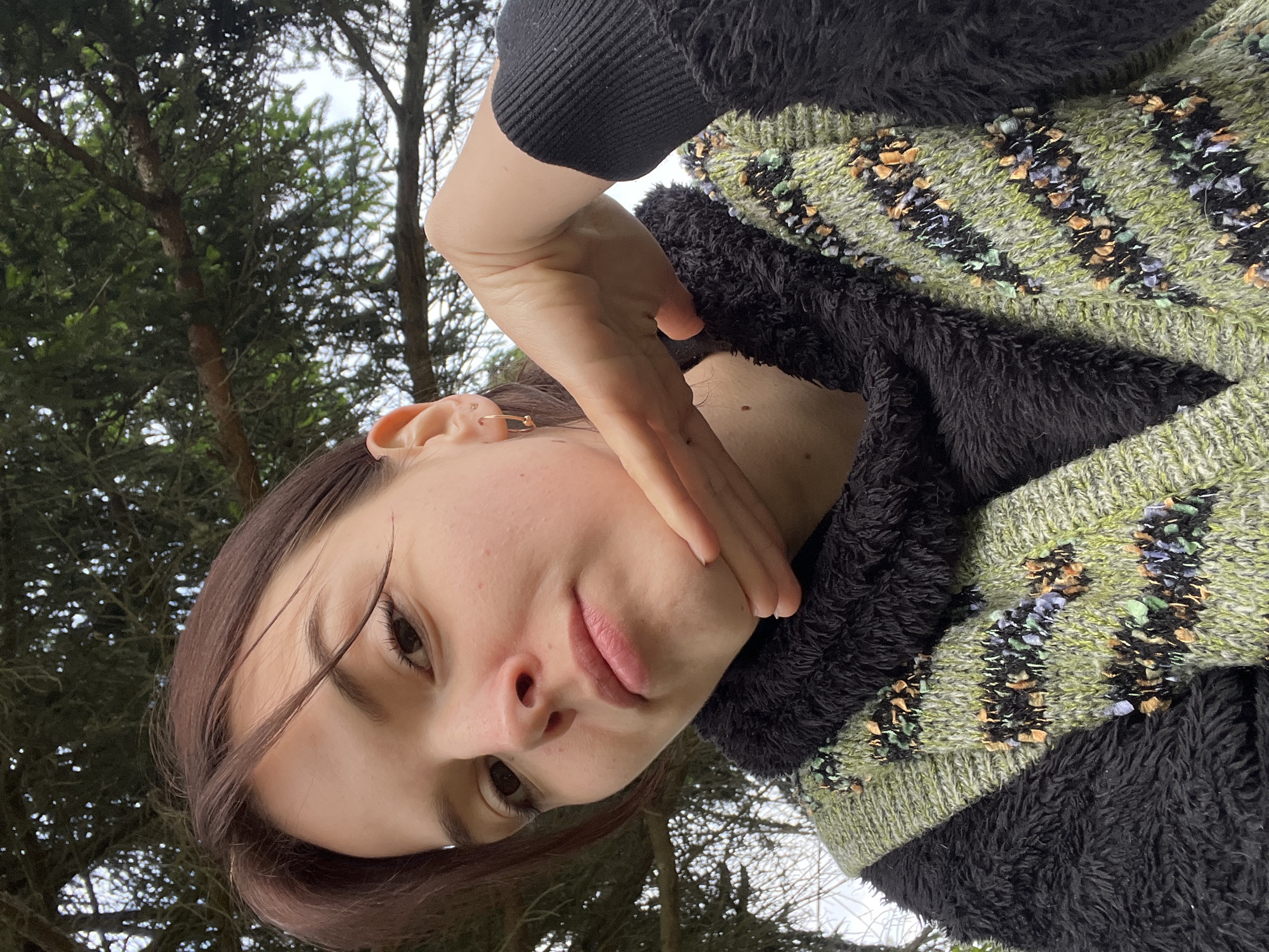A young woman is pictured outdoors in what appears to be a forested area near Mshinskaya, Russia. She has light skin, brown hair, and is wearing a black fuzzy jacket over a green and black sweater with decorative stripes. Her right hand is partially covering her face. The background shows trees against a cloudy sky. The photo is taken from a low angle, looking up at the subject.