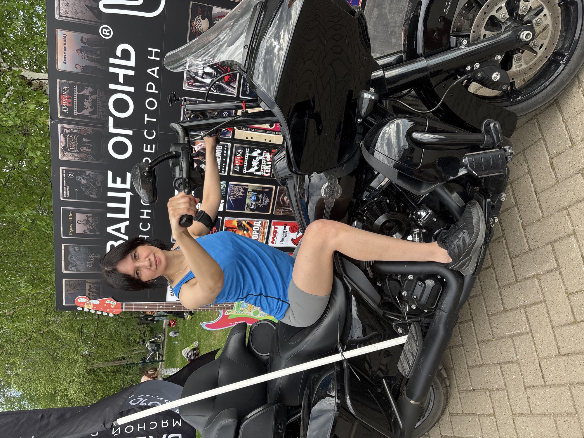 Here is a detailed description of the image:
**Scene**: The image depicts a woman sitting on a black Harley-Davidson motorcycle, positioned on a paved surface, possibly a street or plaza.
**People**: A woman with short, dark hair, wearing a blue tank top and grey shorts, is sitting on the motorcycle. She is smiling and looking towards the camera.
**Objects**:
* A black Harley-Davidson motorcycle.
* A backdrop with a sign that reads "НАШЕ ОГОНЬ CHO РЕСТОРАН" along with numerous cassette and CD covers of rock bands. A painted guitar is also visible on the board.
* A flag with lettering (partially obscured).
* Paved surface made of rectangular blocks.
**Environment**: The setting appears to be outdoors, with greenery and people in the background suggesting a public space. The lighting appears to be natural daylight.
**Location**: The scene is in the city of Unknown, Unknown.