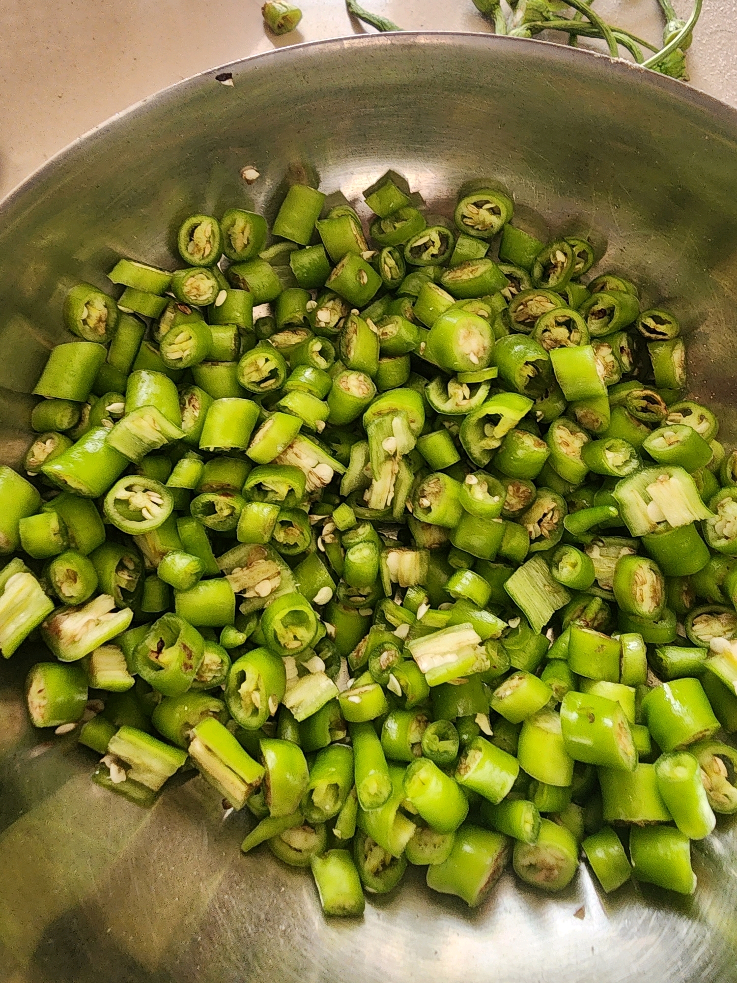 In a metal bowl, a generous portion of chopped green chili peppers are piled high. The peppers are cut into uniform, bite-sized pieces, revealing their bright green exteriors and pale yellow, seedy interiors. Some pieces show the stark white of the chili pith. The bowl itself appears to be made of stainless steel and rests on a light-colored, slightly textured surface, possibly a countertop. In the background, the tips of more green chili peppers, still attached to their stems, are visible, suggesting they were recently prepared for chopping. The lighting suggests an indoor setting, possibly a kitchen in Mahesana, India, given the culinary context of chili peppers. There are no people or discernible actions taking place; the image focuses solely on the prepared ingredients.