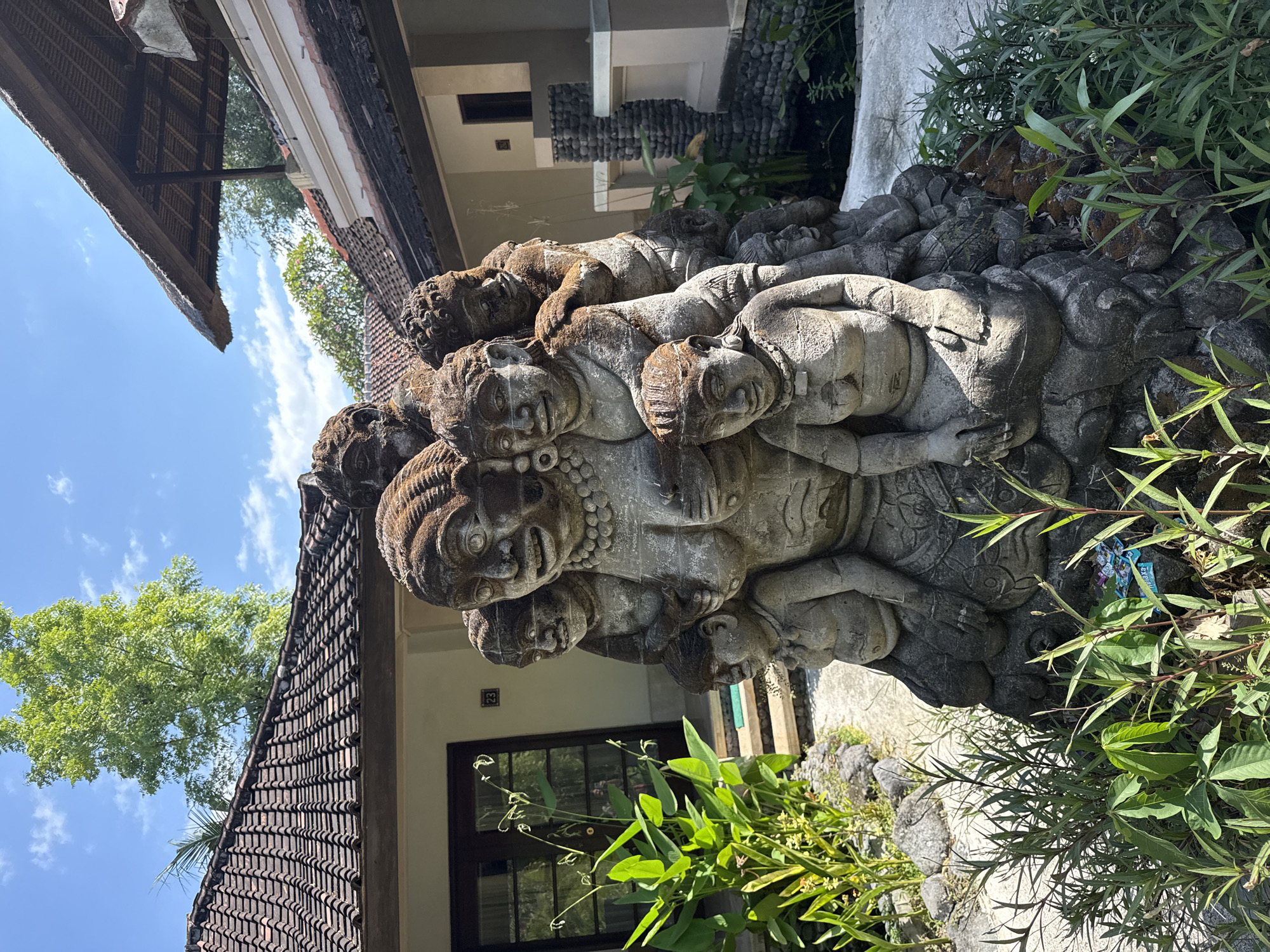 The image shows a stone sculpture of a family in an outdoor setting. The sculpture depicts a large central figure, presumably a mother, surrounded by several smaller figures that appear to be children. The figures are carved in a somewhat stylized, traditional Balinese manner and show signs of weathering, with moss and discoloration visible on the stone. The sculpture is placed in front of a building with a tiled roof and a traditional Balinese thatched roof structure nearby. Lush green vegetation, including tall plants and smaller bushes, surrounds the sculpture and the building. The background shows a clear blue sky with some white clouds. The scene appears to be in a tropical or subtropical environment, consistent with locations like Kubu, Indonesia. There are no people actively engaged in any actions in the scene; the focus is on the static sculpture and its surroundings.