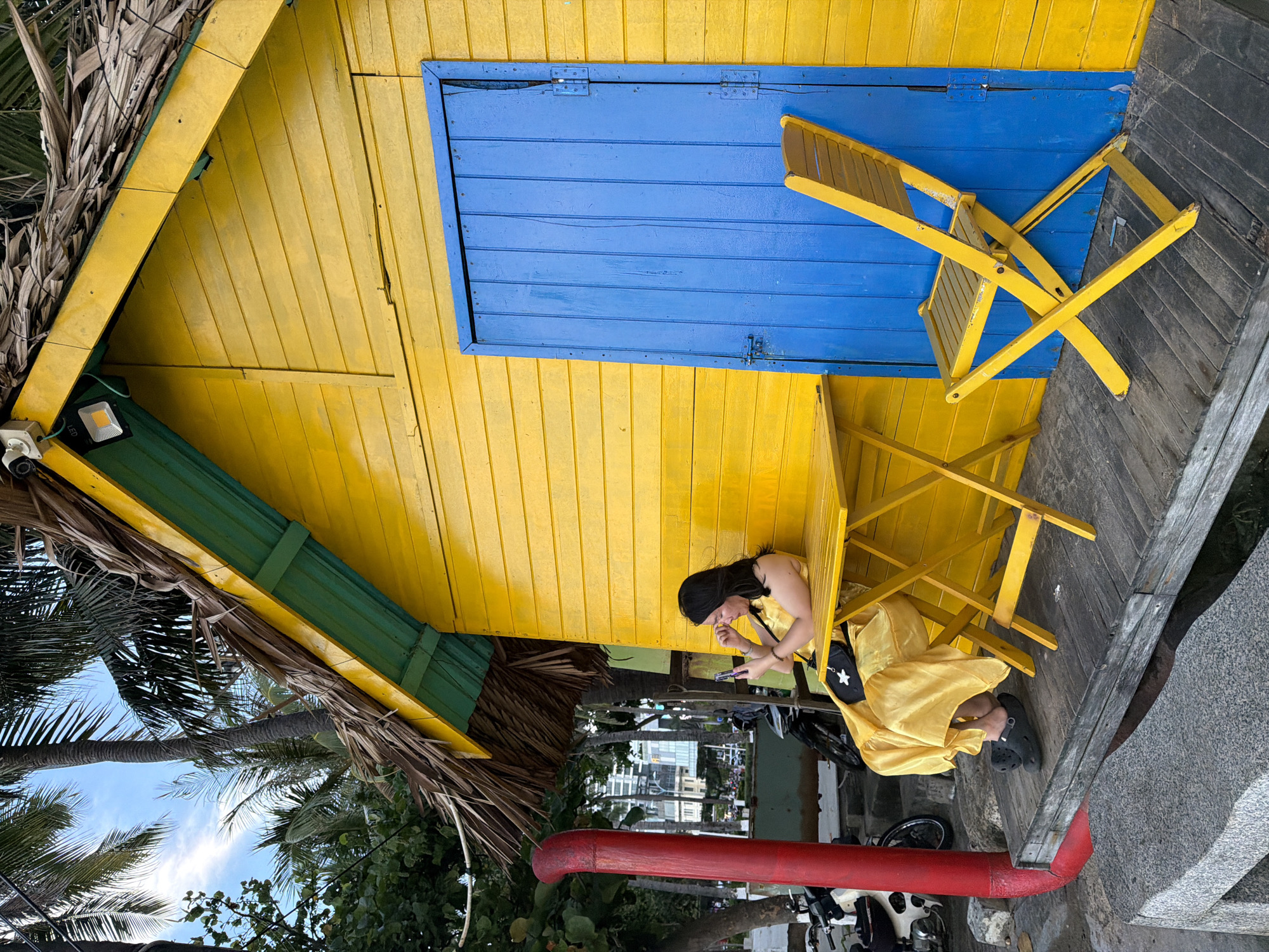 A woman is seated at a yellow table outside a vibrant yellow wooden building with a thatched roof. She is wearing a flowing yellow dress and looking at her phone. A bright blue door is on the side of the building. To the right of the door, a yellow folding chair is placed on a wooden deck. In the background, palm trees and buildings can be seen. The scene appears to be located in Nha Trang Ward, Vietnam, suggested by the architectural style and vegetation.