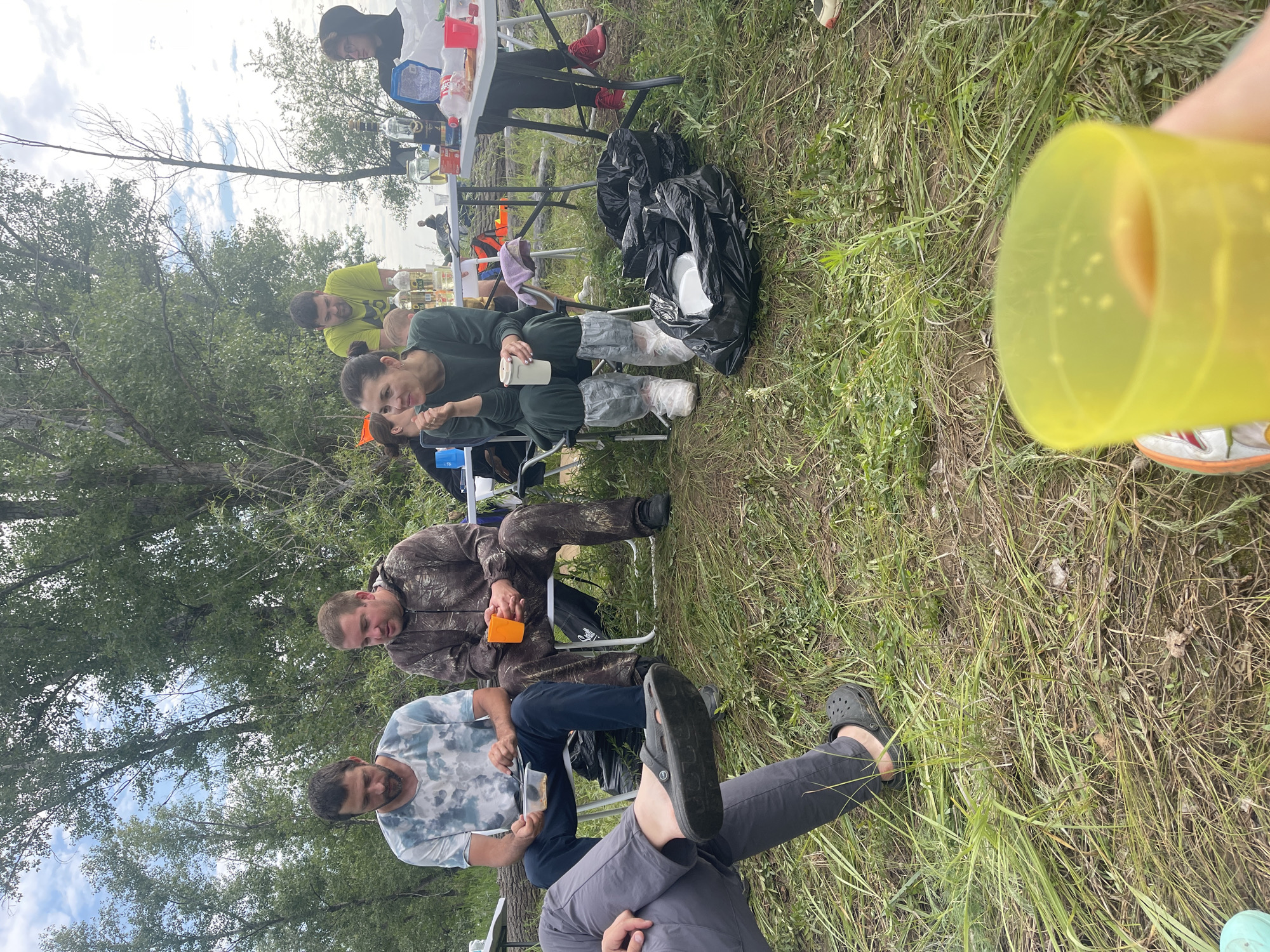 A group of adults is gathered outdoors, likely in a park or recreational area near Nezhinka, Russia, for a casual meal. They are seated around foldable tables laden with food and drinks, with the backdrop of a wooded area and a cloudy sky.
In the foreground, a person wearing grey pants and black Crocs is partially visible, holding a yellow plastic cup.
To their left, a man in a tie-dye t-shirt and dark blue pants sits in a white chair, holding a white bowl. Beside him, another man in camouflage clothing sits in a white chair, holding an orange plastic cup.
Further back, a woman in a dark green outfit is seated, holding a white cup. A person in a bright yellow t-shirt stands behind a table. On the right, a person in black pants and red sneakers is seated.
Several black trash bags are scattered on the ground, some containing disposable plates. The environment is grassy with tall trees and bushes. The overall atmosphere suggests a relaxed outdoor gathering.