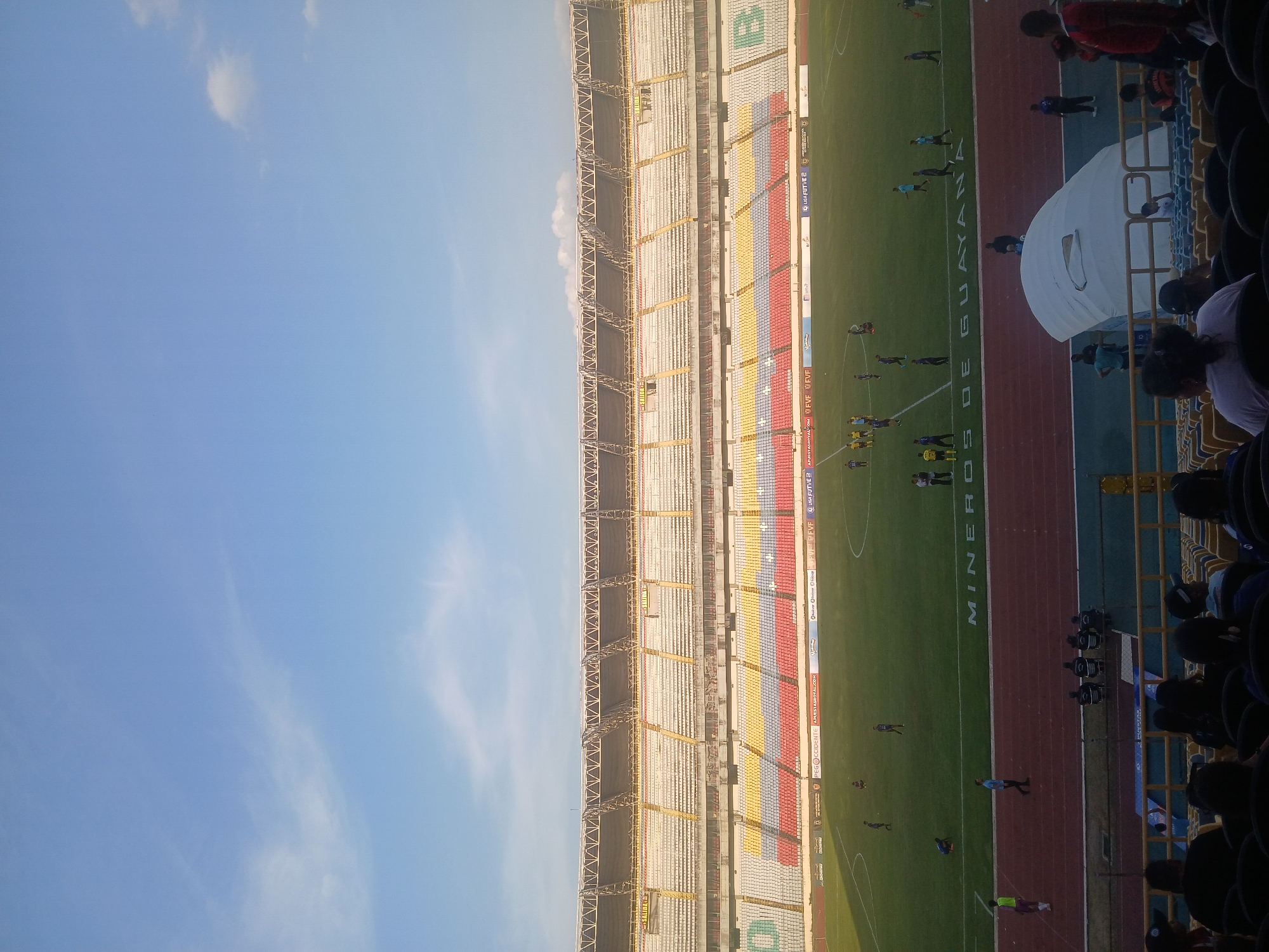 The image depicts Estadio CTE Cachamay in Ciudad Guayana, Venezuela. It shows a view of the stadium with visible seating sections, a football field, and a running track. There are people on the field and in the stands. The sky is visible in the background.