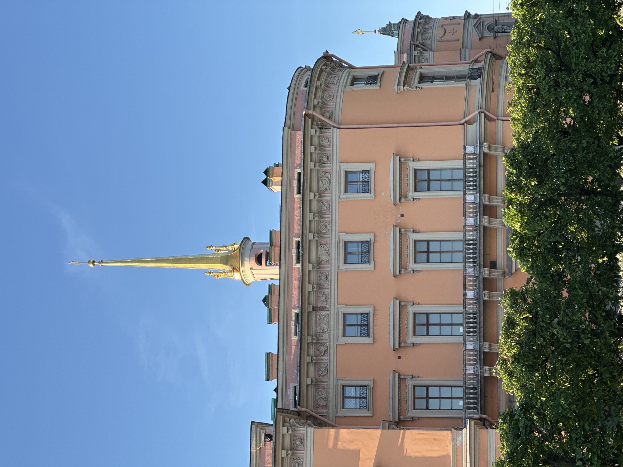 The image, oriented correctly, showcases a distinctive and ornate architectural structure against a clear, bright blue sky. The main subject is a large building, predominantly painted in a warm salmon pink or peach color, exhibiting a rich Baroque or Rococo style.
The building's facade is highly decorative, featuring numerous rectangular windows with prominent white frames. Some windows are protected by elaborate metal grilles. Horizontal friezes adorned with intricate sculptural details and motifs run across different levels of the building. Sections of the structure display elegant curves, cornices, and balconies with decorative balustrades. Visible brown drainpipes are integrated into the design.
A prominent golden spire rises from a circular tower section of the building, tapering upwards to a pointed finial. Smaller decorative elements and turrets are visible along the roofline. On the right side, a substantial tree with vibrant green foliage partially obscures the building, adding a natural element to the scene.
The bright sunlight and cloudless sky indicate a clear daytime setting. There are no people or specific activities depicted. No discernible text is visible in the image. The architectural style, color palette, and the golden spire, combined with the location context, strongly suggest the building is Mikhailovsky Castle (also known as St. Michael's Castle or Engineers' Castle) in Saint Petersburg, Russia.