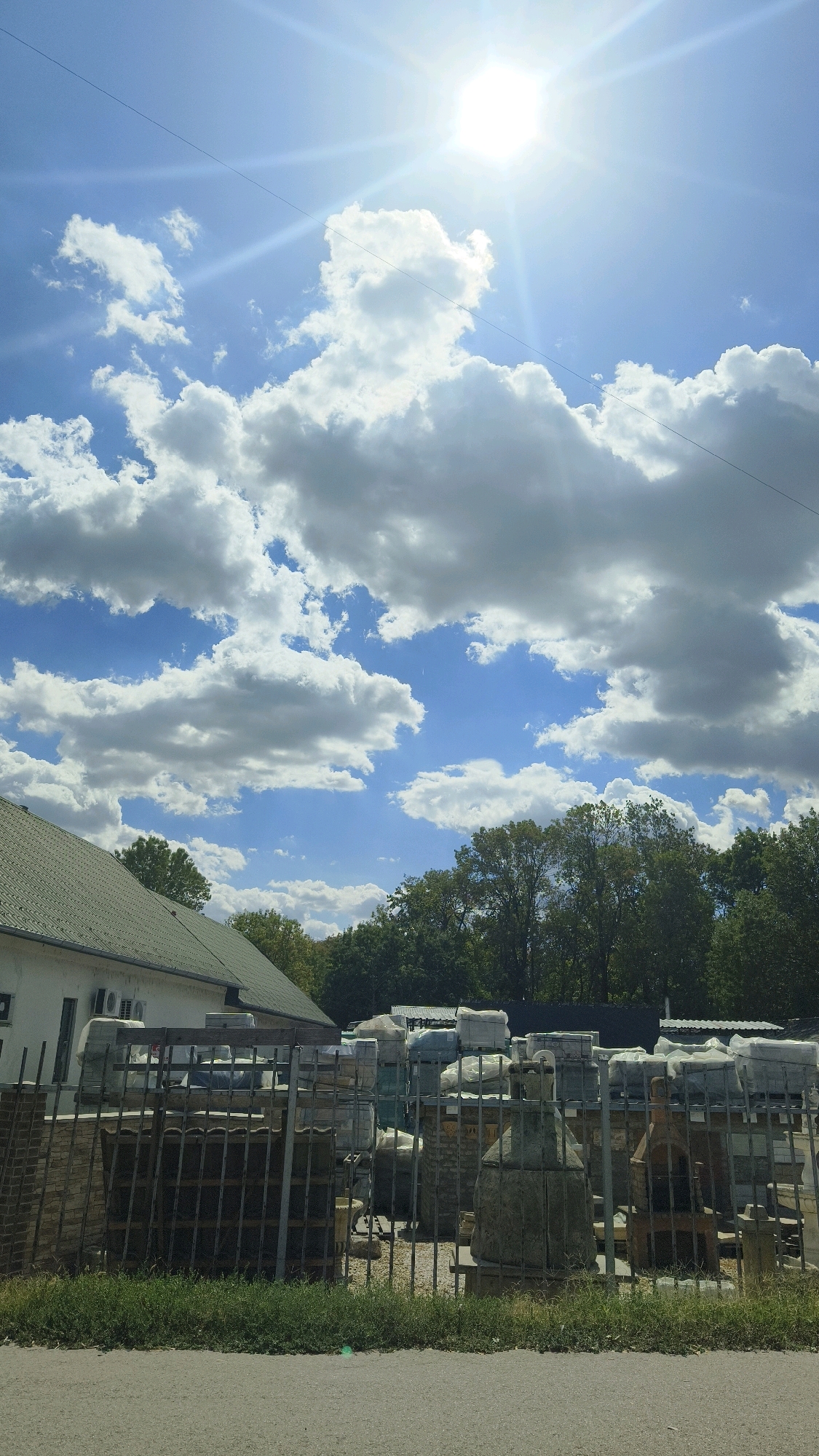 The image captures an outdoor scene, likely a storage or sales yard, under a bright, sunny sky with scattered cumulus clouds. The sun is prominently positioned in the upper right quadrant, casting bright rays. In the foreground, a metal fence encloses a collection of stacked materials, possibly building supplies or garden ornaments, some of which are covered in plastic sheeting. Behind the fence, a white building with a green roof is visible on the left, and a dense line of trees forms the background. The scene suggests a commercial area within the city of Szeged, Hungary, given the context. There are no people or active interactions depicted.
