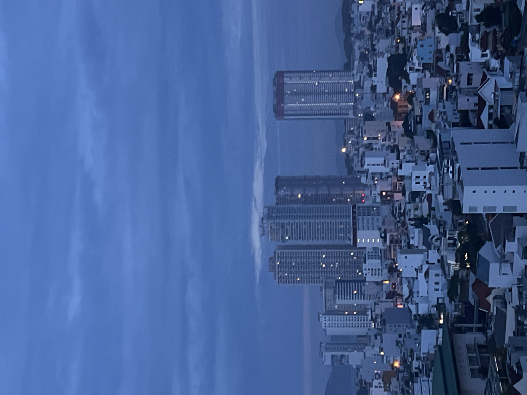 The image captures an expansive, high-angle view of a coastal city at dusk or night, located in what appears to be Bac Nha Trang Ward, Vietnam. The sky, occupying the upper left portion of the frame (when viewed with correct orientation), is a deep, muted blue with soft, diffuse clouds, suggesting the transition from twilight to full night.
The urban landscape dominates the right side of the image, showcasing a densely packed array of buildings. Several prominent high-rise towers, characterized by their rectangular forms and numerous illuminated windows, stand out against the darker backdrop. These larger structures are surrounded by and interspersed with a multitude of smaller residential and commercial buildings, many of which also display glowing windows or streetlights, creating a scattered pattern of warm light across the cityscape. The architectural style appears varied, encompassing modern high-rises and more traditional, lower-rise constructions.
A dark body of water, likely a bay or the ocean, stretches across the mid-ground, separating the city from the distant horizon. Faint outlines of land or mountains are visible in the far distance, blending with the low light conditions. No people are visible, and no specific activity or event is depicted beyond the general illumination of the city at night. The scene conveys a sense of urban tranquility and vastness during the evening hours. There is no visible text in the image.