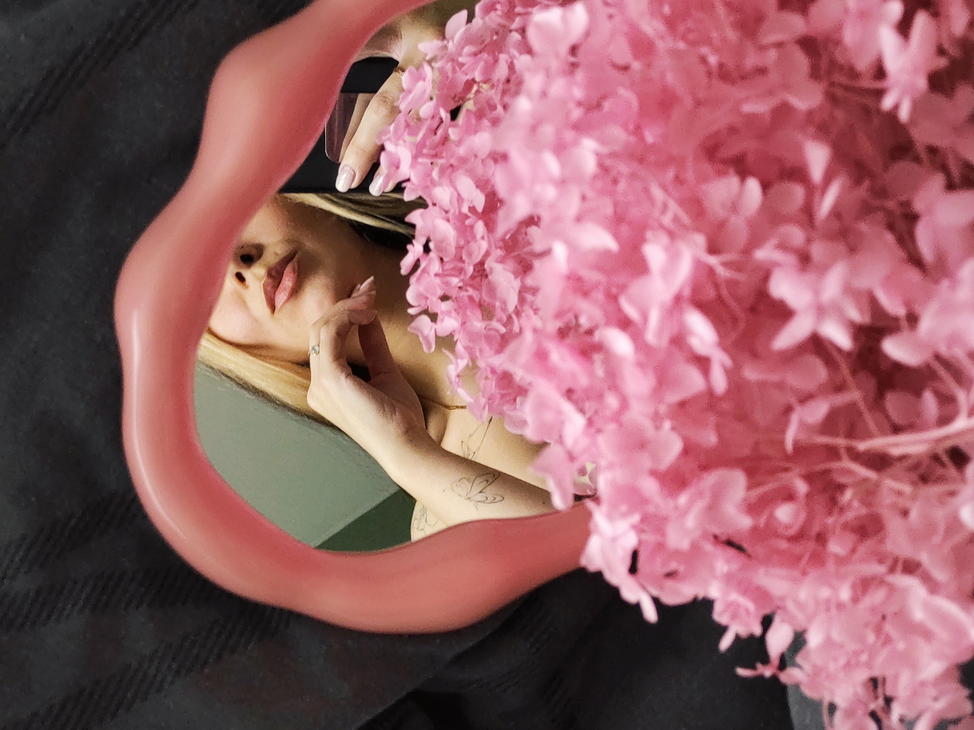 This image features a partial reflection of a person, likely female, captured within an irregularly shaped, wavy pink mirror. The mirror's organic frame curves around the subject. The right side of the composition is largely filled with a dense cluster of light pink, fluffy flowers, possibly dried hydrangeas, which softly obscure parts of the reflection and the background.
Through the mirror, the subject's face is seen in profile, with her blonde hair partially visible. Her lips are slightly parted, conveying a relaxed or posed expression. Her right hand, featuring a ring on one finger and manicured nails, rests gently near her neck. A small butterfly tattoo is visible on her forearm. Her left hand, also with manicured nails, is seen holding a dark, metallic object that could be a phone or a small case. A delicate gold chain necklace is visible around her neck. The background within the mirror's reflection is a plain, light green surface.
Outside the mirror's frame, and partially around the reflected image, a dark, textured fabric, likely a blanket or clothing, creates a contrasting backdrop. The overall lighting is soft and indirect, suggesting an indoor setting. The image appears to be a carefully composed self-portrait or artistic shot, emphasizing softness, intimacy, and a subtle allure. No visible text is present in the image. The location context is Chaikovsky, Russia.