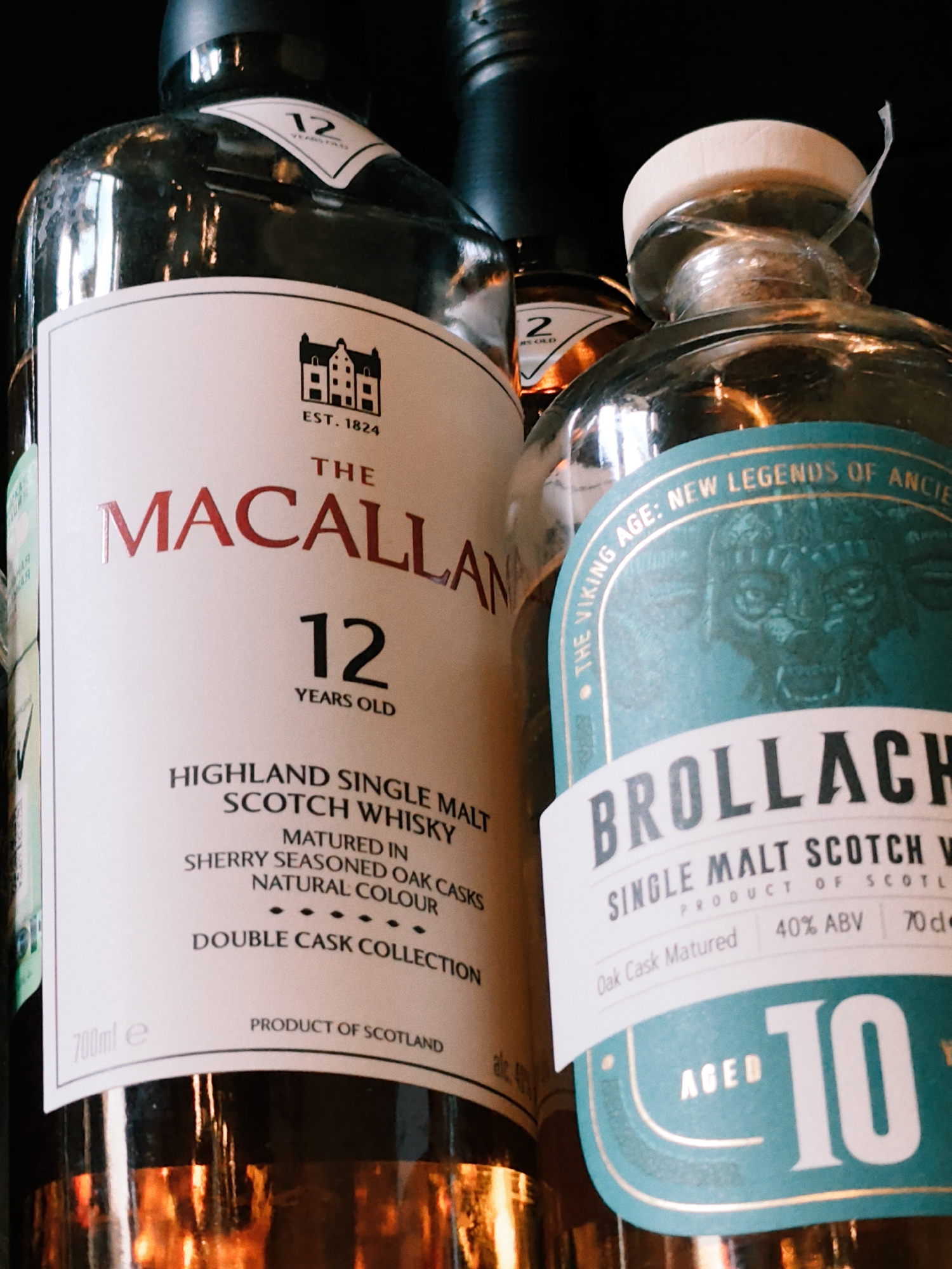 The image shows two bottles of Scotch whisky. The bottle on the left is The Macallan 12 Years Old, a Highland Single Malt Scotch Whisky matured in sherry seasoned oak casks. It is 700ml and a product of Scotland. The bottle on the right is Brollach, a Single Malt Scotch Whisky with "The Viking Age: New Legends of Ancient" written in gold on a teal label. It states "Aged 10" and "40% ABV 70cl". The bottles are presented against a dark background, with reflections on the glass indicating an indoor setting. There are no people present in the image. The location of the image is not specified within the image itself and cannot be determined from the visual information provided.