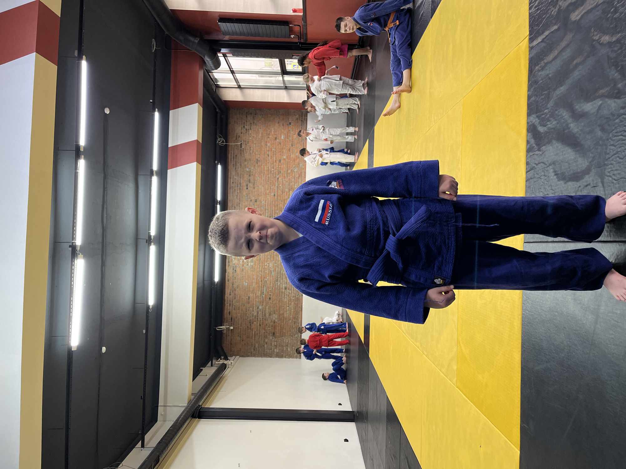 A young male, appearing to be around 10-12 years old, stands center frame, facing the camera with a stern expression. He is wearing a dark blue judo uniform with a white belt tied around his waist and a Russian flag patch on his left shoulder. The background shows a martial arts training facility with yellow and black mats. Several other children in judo uniforms are visible in the background, some are training, some are observing. The room has exposed brick walls, fluorescent lighting, and a high ceiling with visible ductwork. The overall impression is of a judo practice session. Given the Russian flag patch on the uniform, this scene is likely taking place in Russia, possibly in a city like Krasnoyarsk where judo is a popular sport.